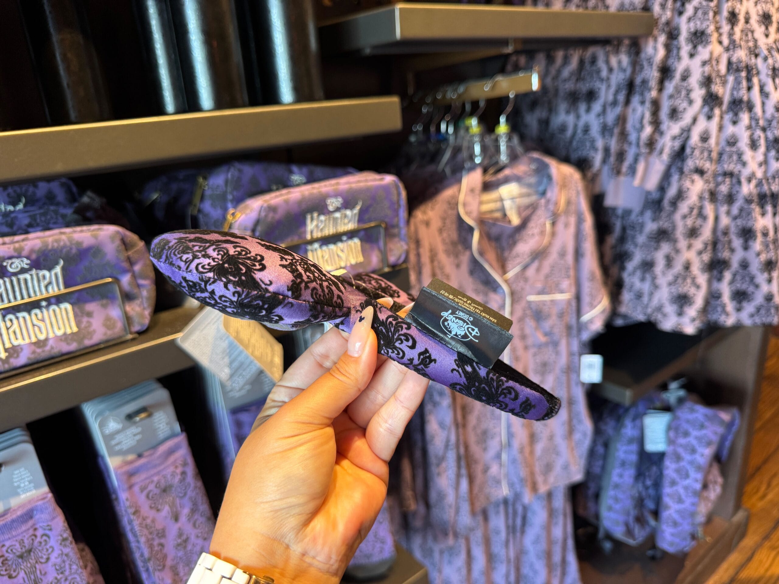 A hand holds a purple Haunted Mansion slipper in a store filled with matching merch and pajamas in the background.