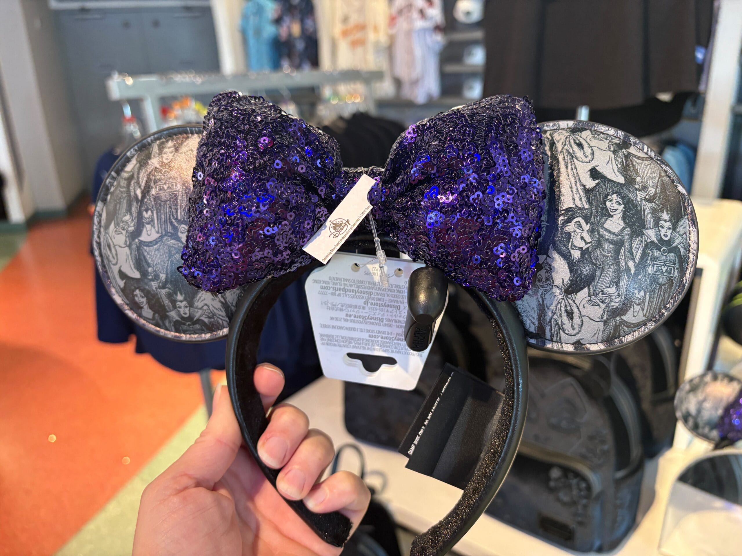 Hand holding Villains ears headband with a purple sequined bow and villain characters, displayed in a store.