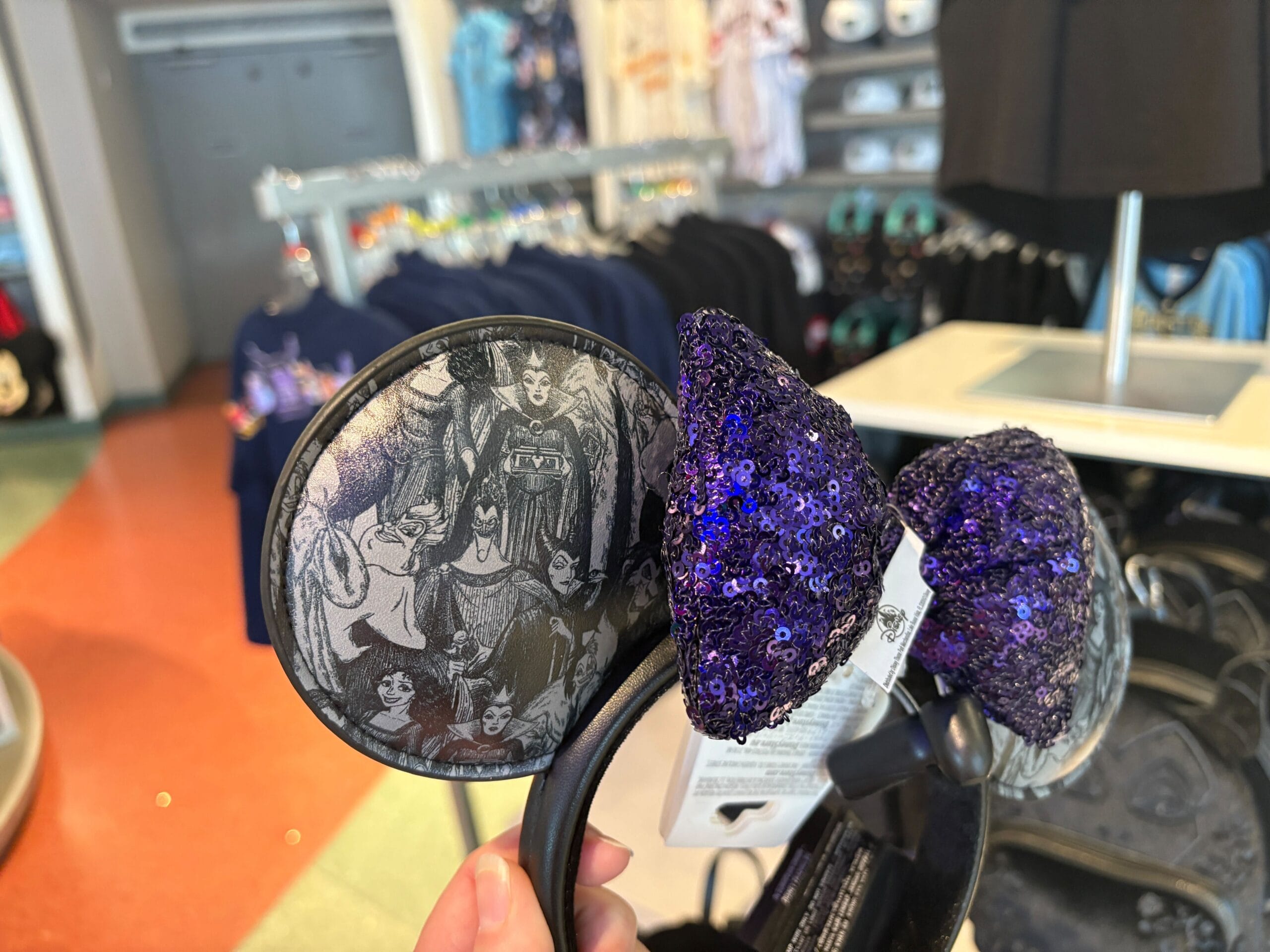 A hand holds Maleficent ears with a purple sequined bow and black-and-white villain print in a Disney store.