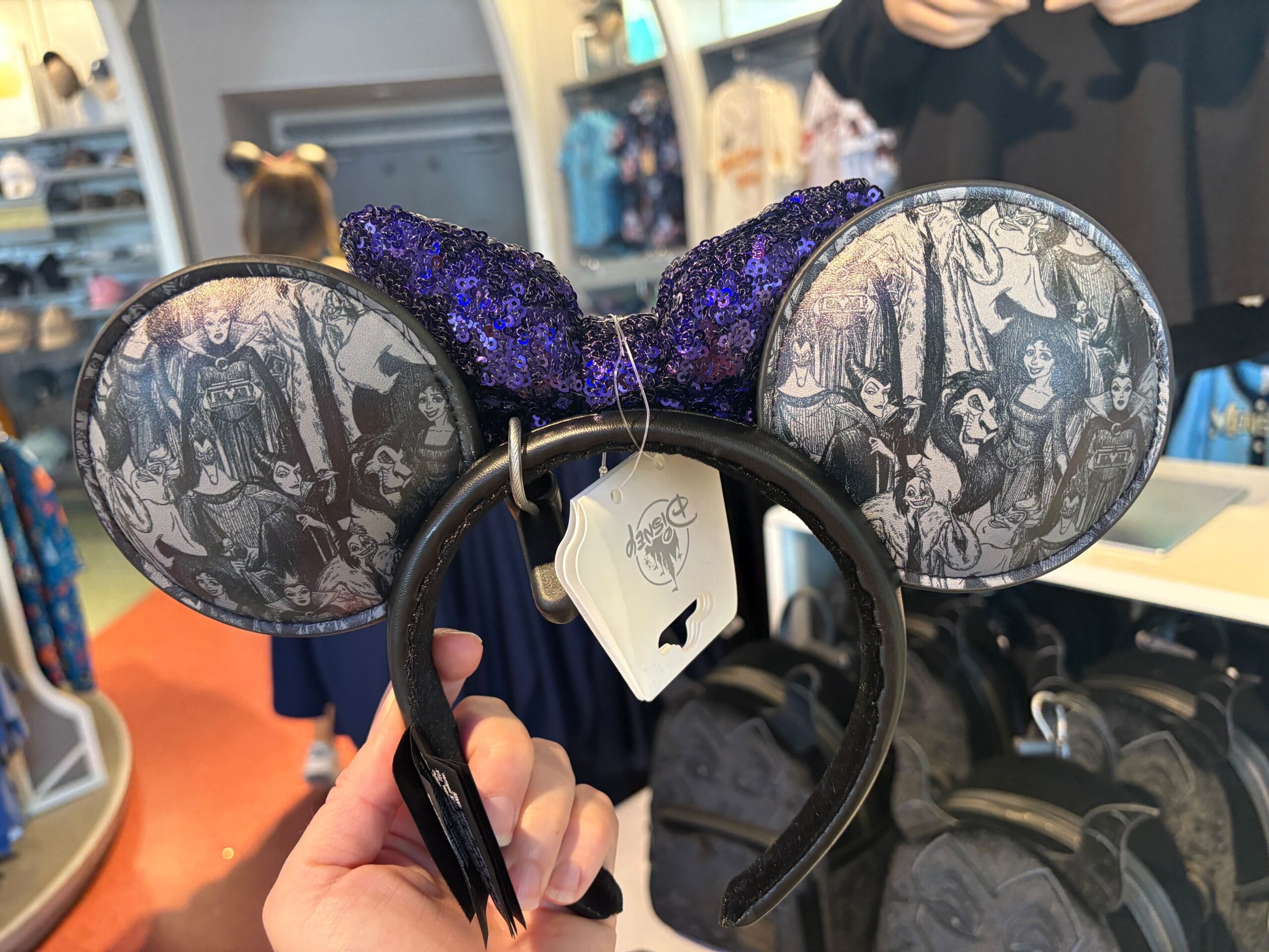 Hand holding Villains ears—black headband with purple sequined bow and Haunted Mansion characters on Mickey Mouse ears.