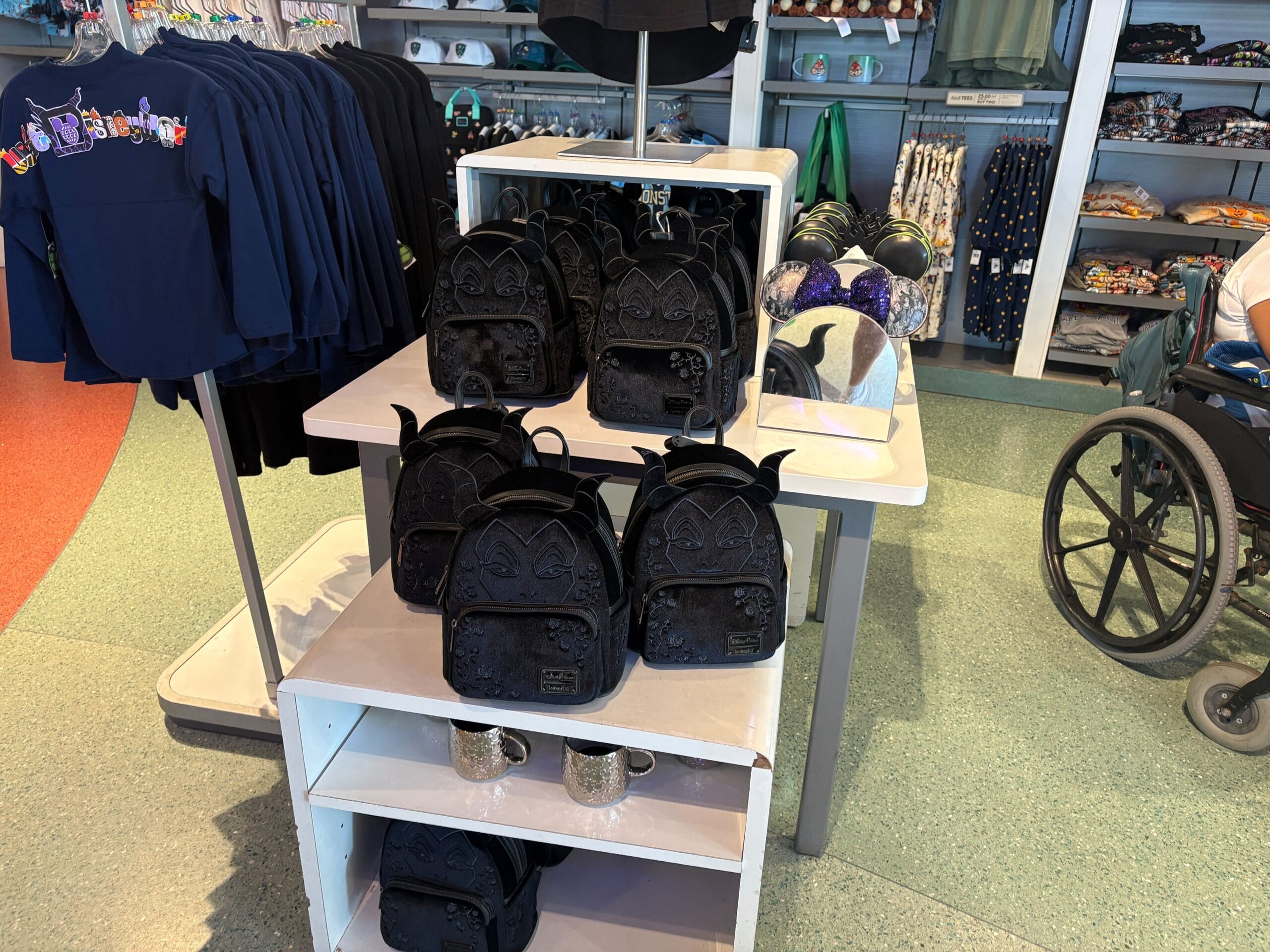 Display of black backpacks with ears on store shelves, surrounded by clothes and hats, near a wheelchair.