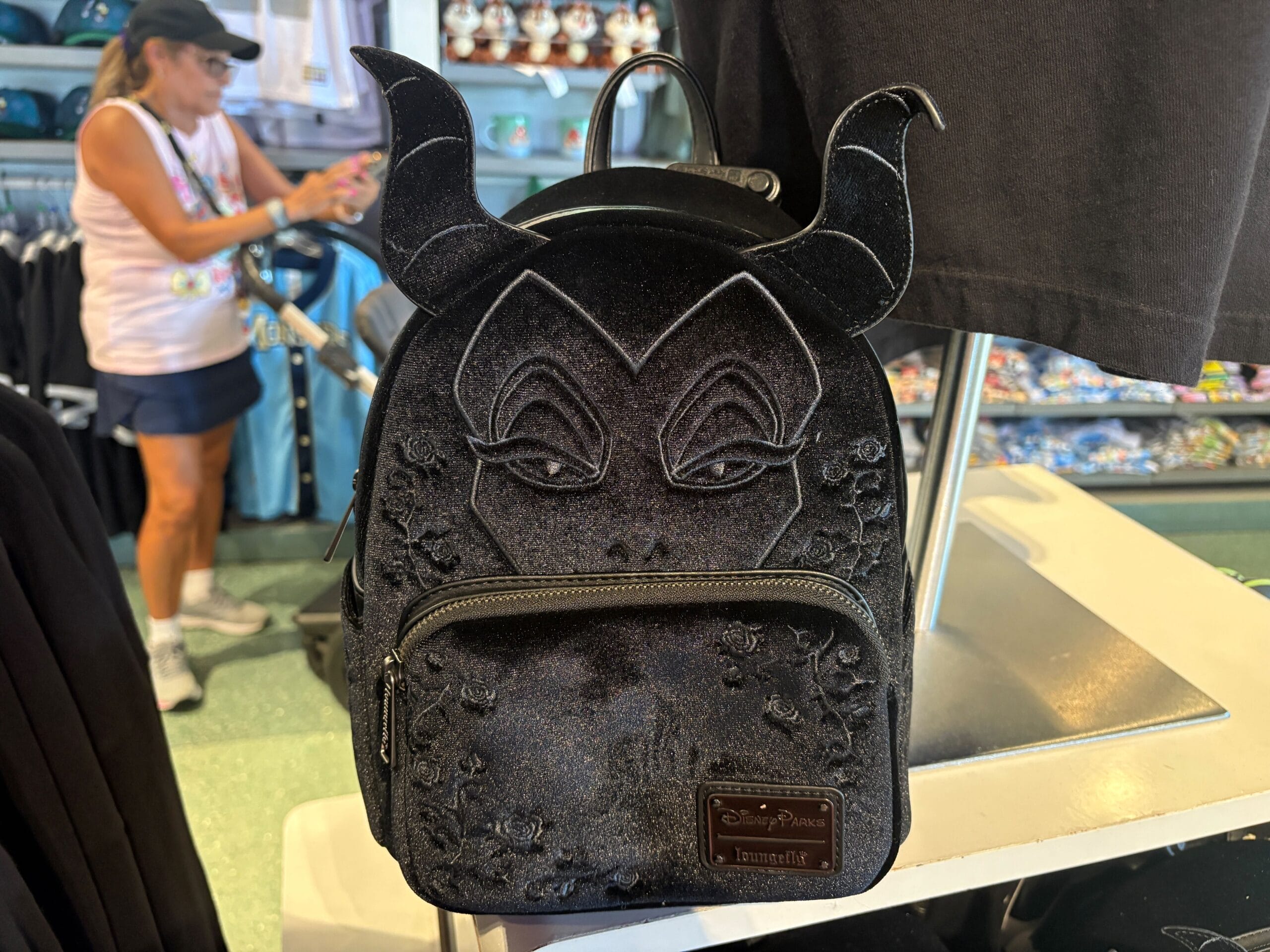 Black backpack with Maleficent's face and horns displayed in a store, with a shopper in the background.