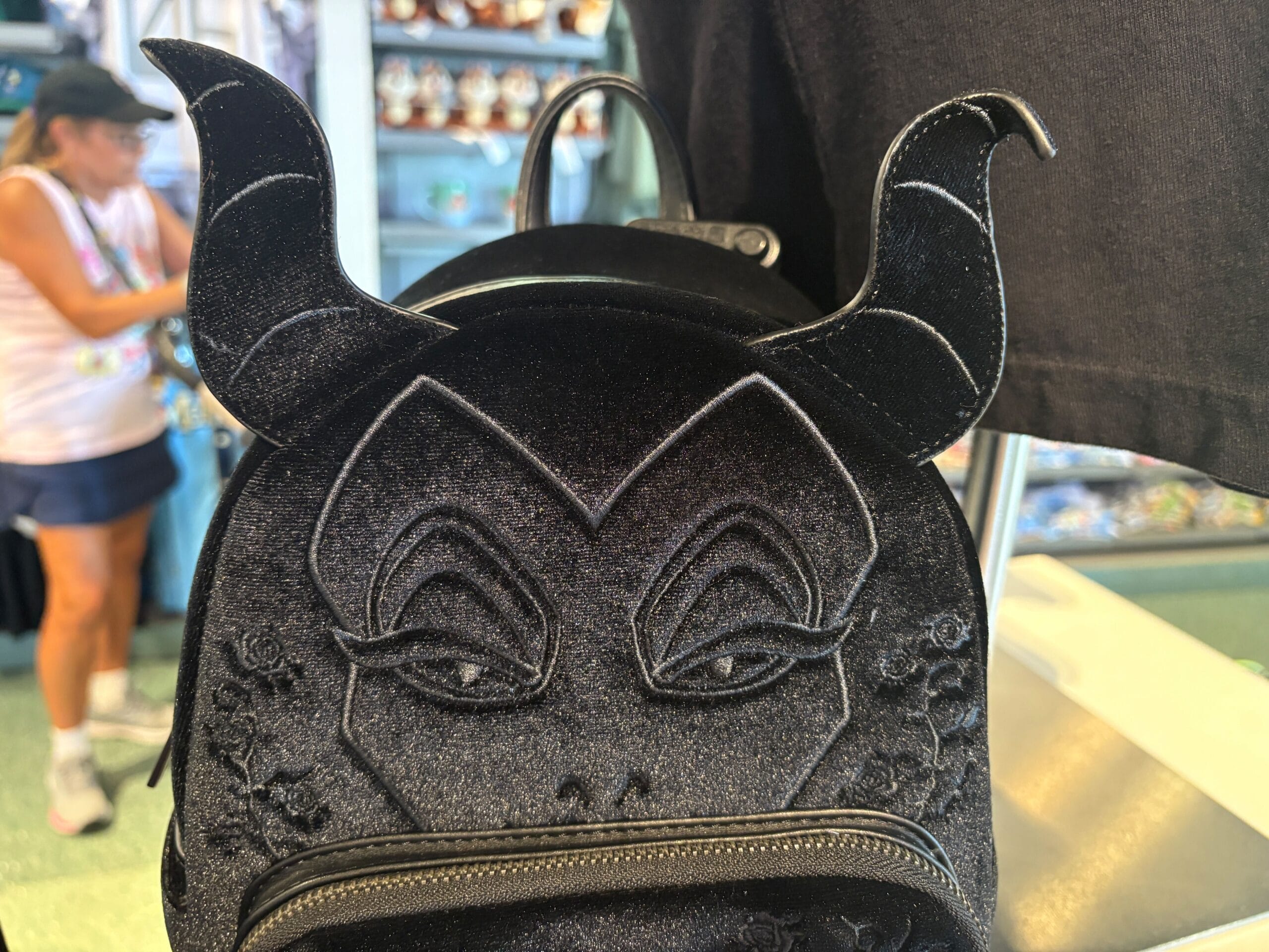 A black backpack with Maleficent's face and horns, displayed in a store with a person in the background.