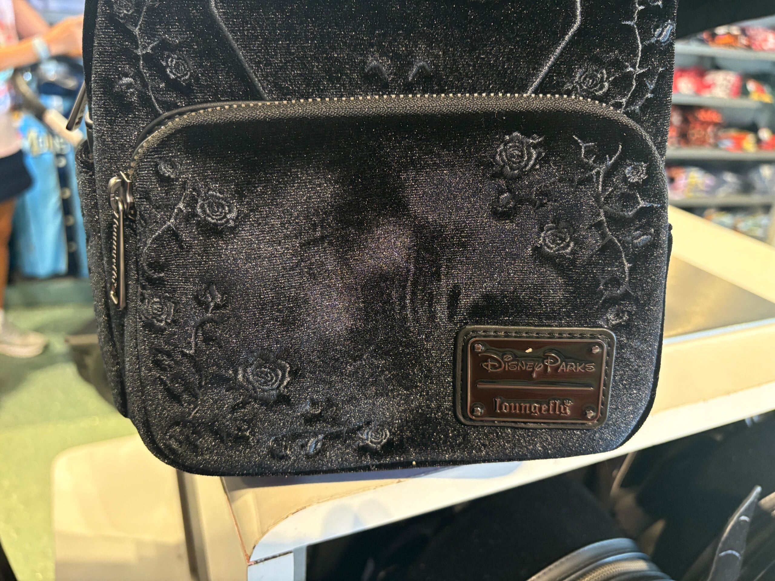 Black velvet Disney Parks Loungefly backpack with embossed roses, displayed on a store shelf.