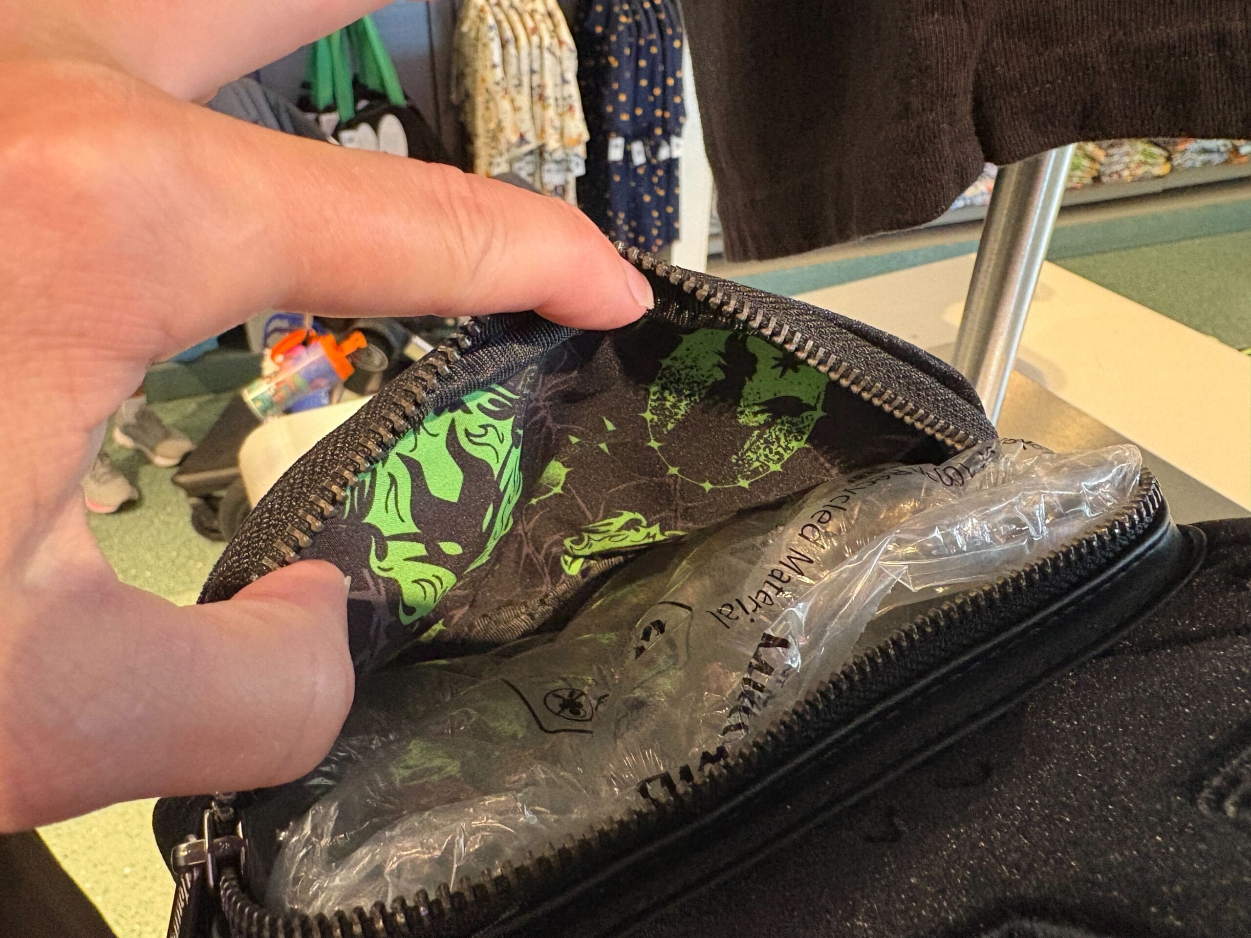 A hand opens a zippered pouch, revealing green-patterned fabric and a clear plastic packing insert inside.