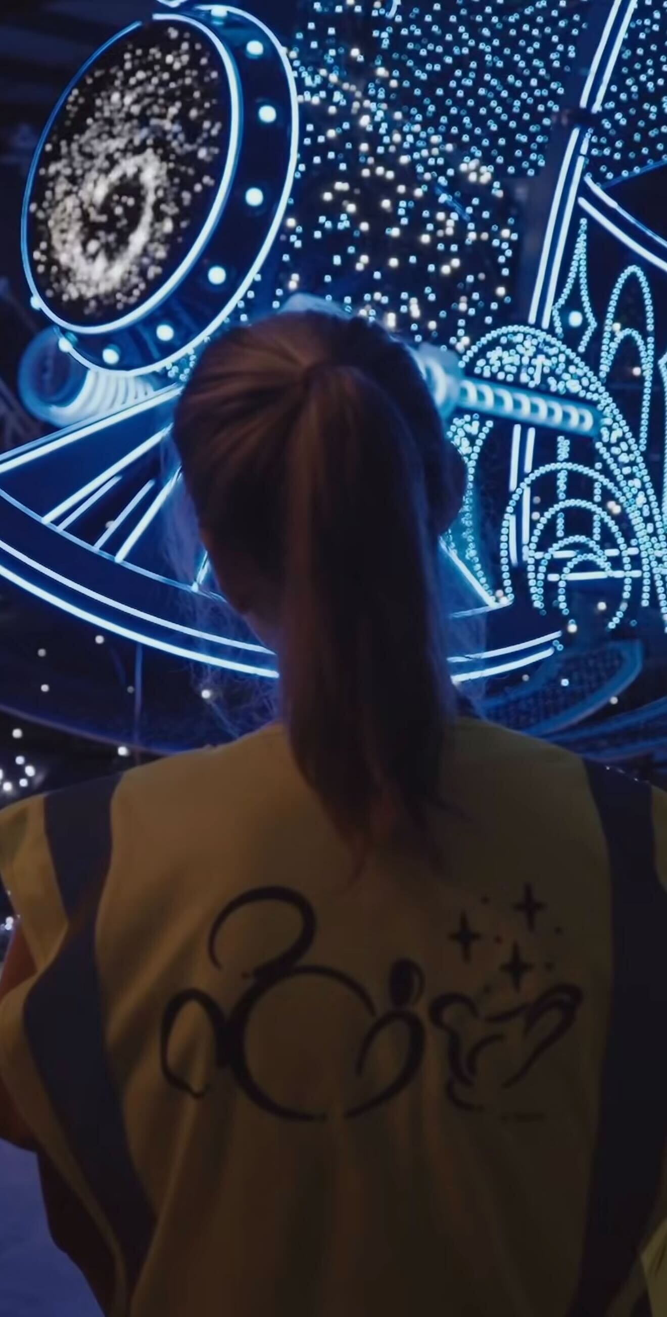 A person in a yellow vest admires a glowing blue galaxy-themed installation, evoking Disney Starlight Parade magic.