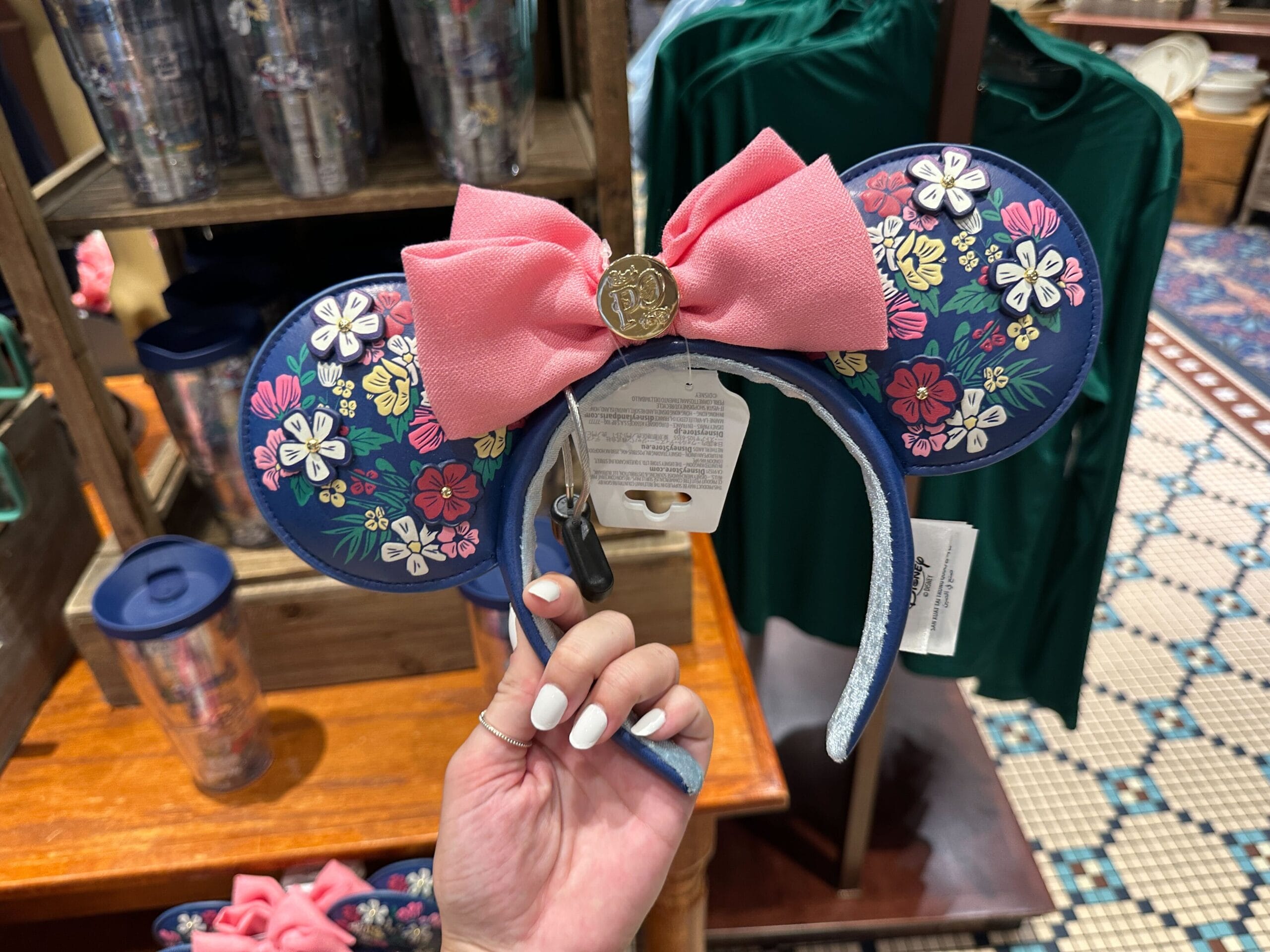 A hand holds floral Minnie Mouse ears with a pink bow and gold emblem, displayed in a store with cups and clothes.