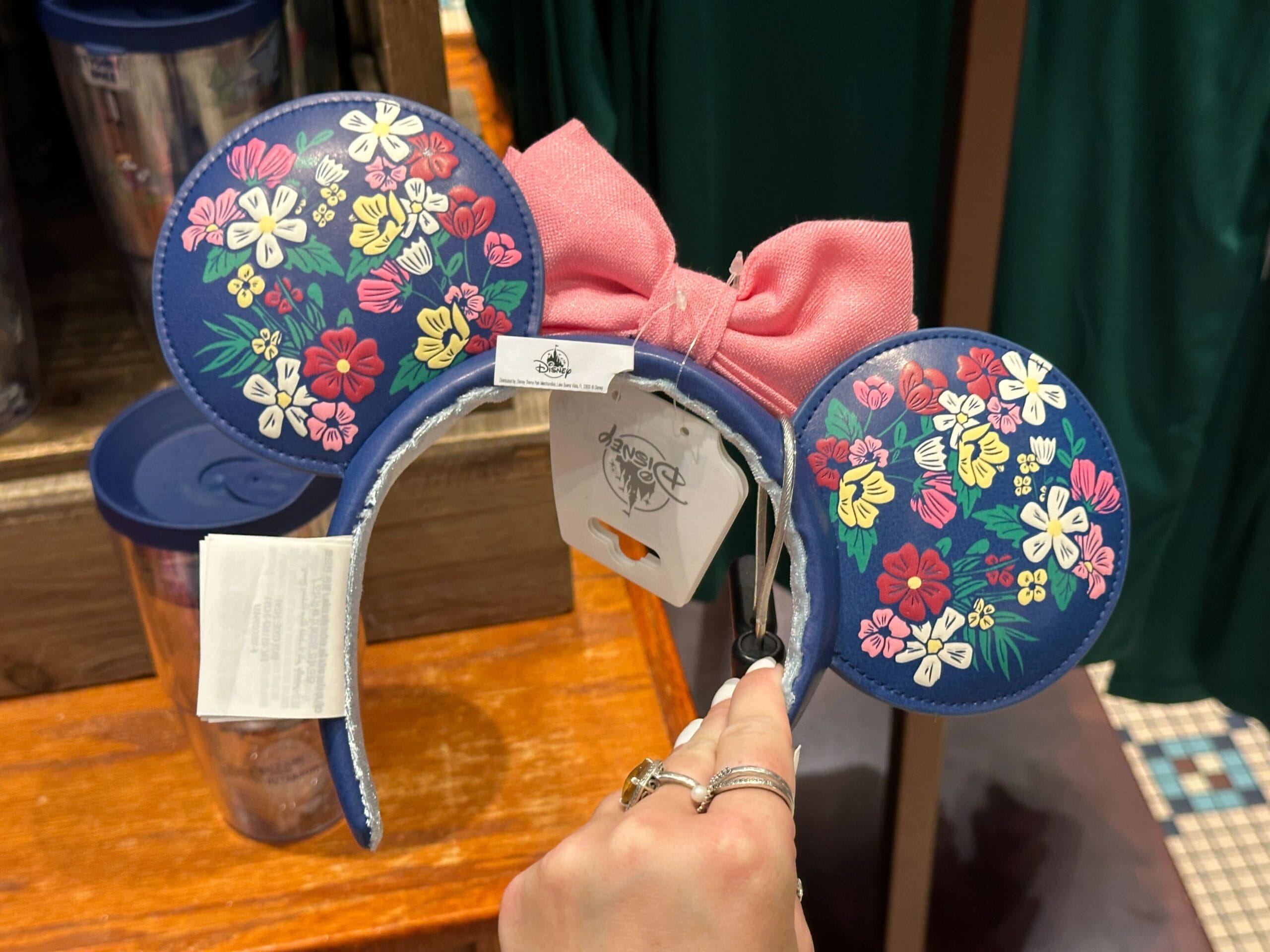 A hand holds Disney mouse ears with floral patterns and a pink bow, displayed in a store.