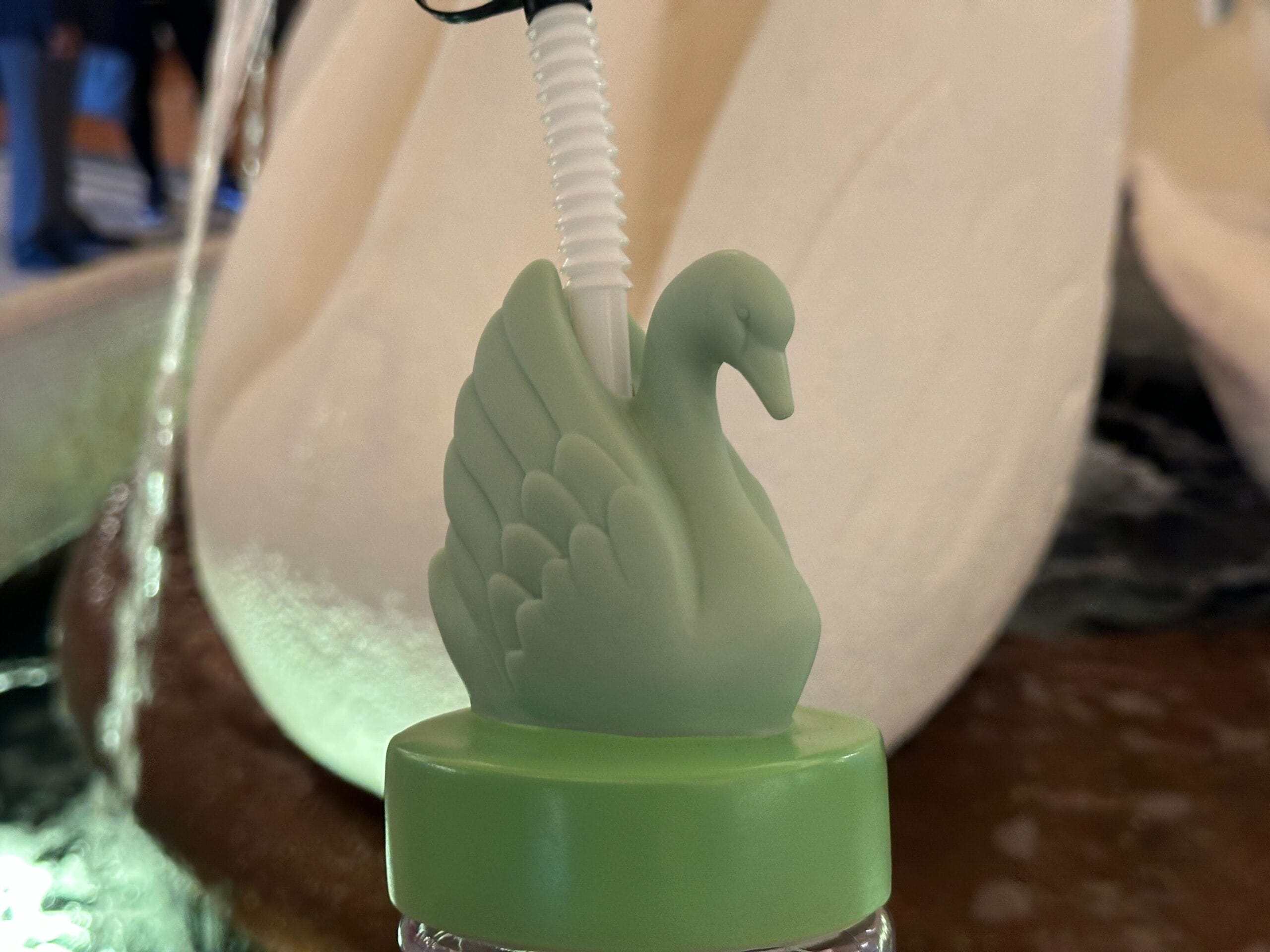 A green swan-shaped Souvenir Sipper with a straw, placed before a white sculpture—perfect for Walt Disney World fans.