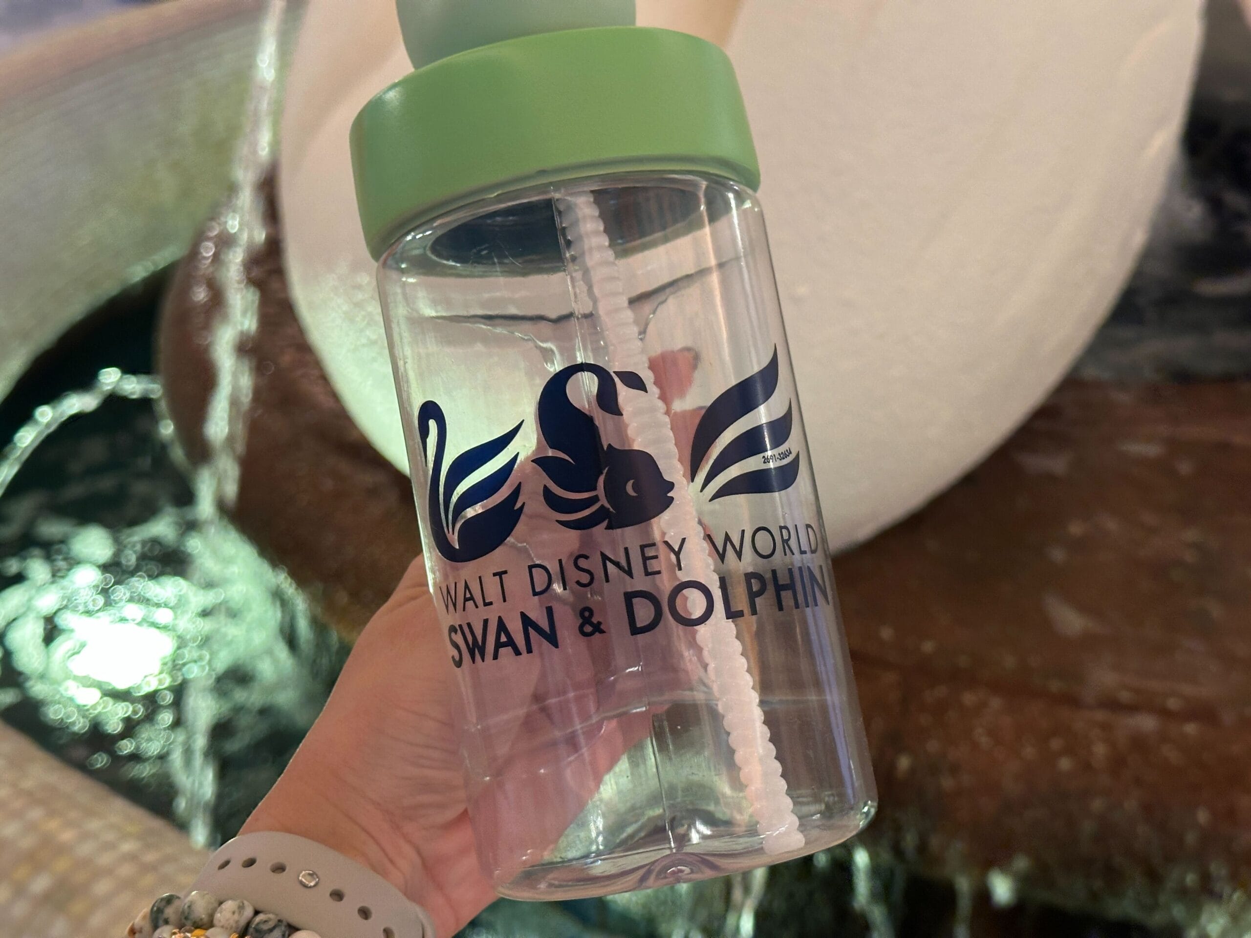 Hand holding a Swan and Dolphin Souvenir Sipper in front of a fountain at Walt Disney World.