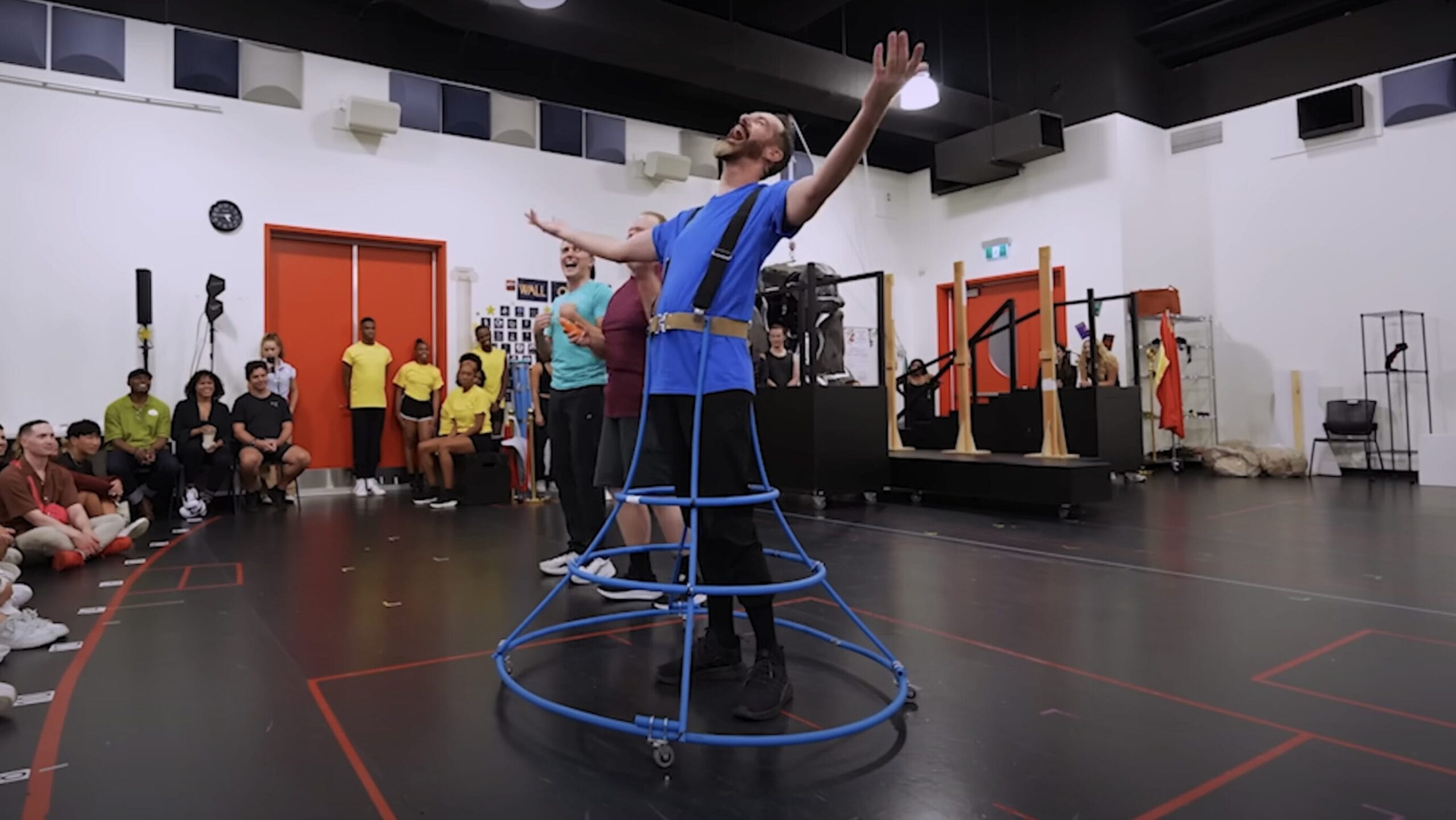 Person in a blue hoop skirt costume poses dramatically during a Hercules Stage Show rehearsal; others watch in awe.