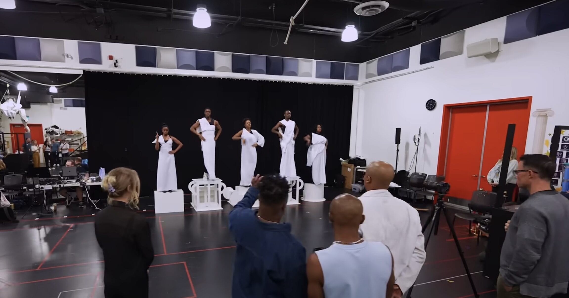Six people in white outfits pose on platforms, recreating a Hercules Stage Show from Disney Cruise Line, as others watch.