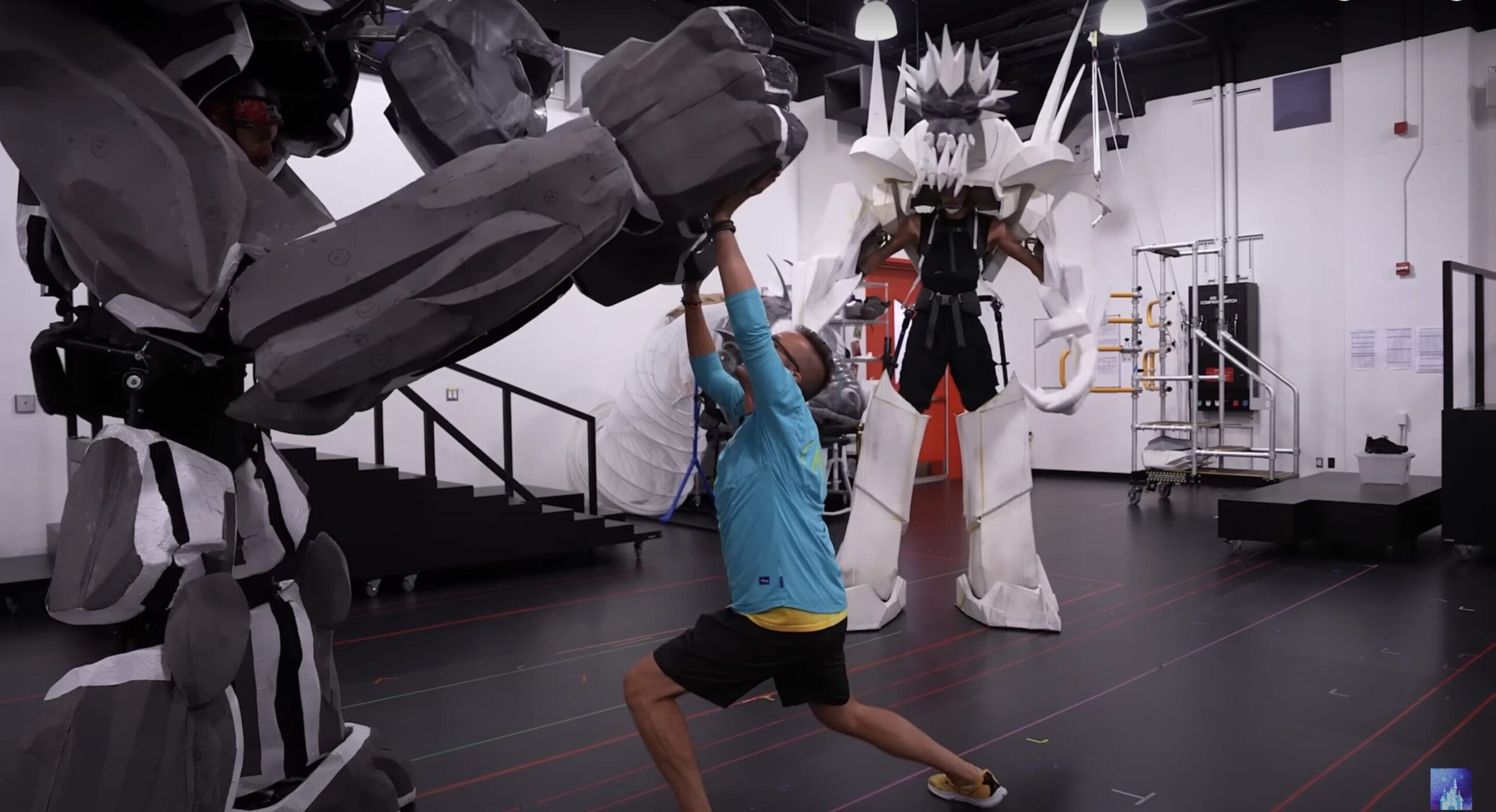 A person poses between two robot costumes, evoking a Zero to Hero vibe in a studio like the Hercules stage show.