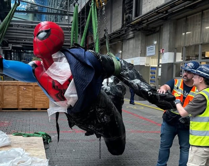 Two workers move a large Stern Character Spider-Man statue suspended by straps in an industrial warehouse.