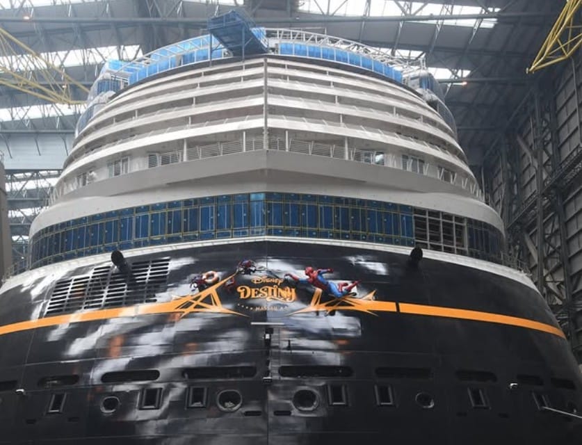 The bow of the Disney Destiny cruise ship under construction, featuring Spider-Man character artwork in a vast indoor shipyard.