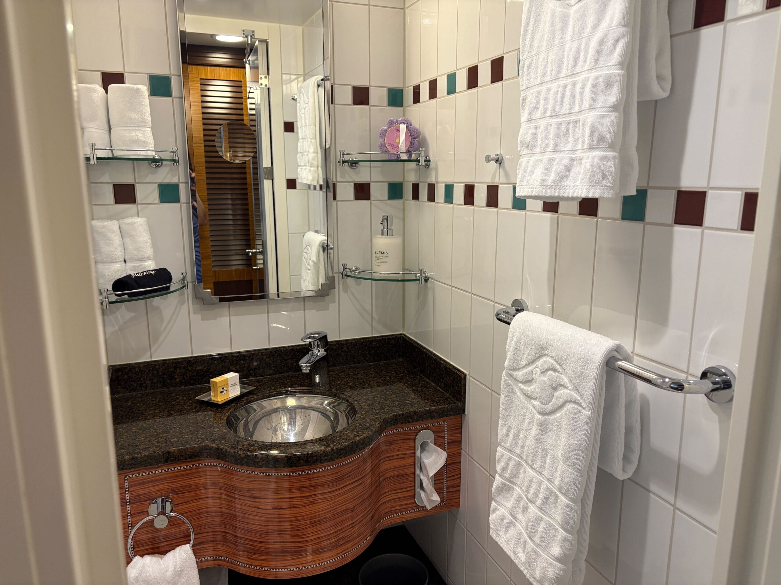 Small bathroom in a Disney Dream Concierge Family Oceanview Stateroom, with white tile, wooden sink counter, shelves, and mirror.