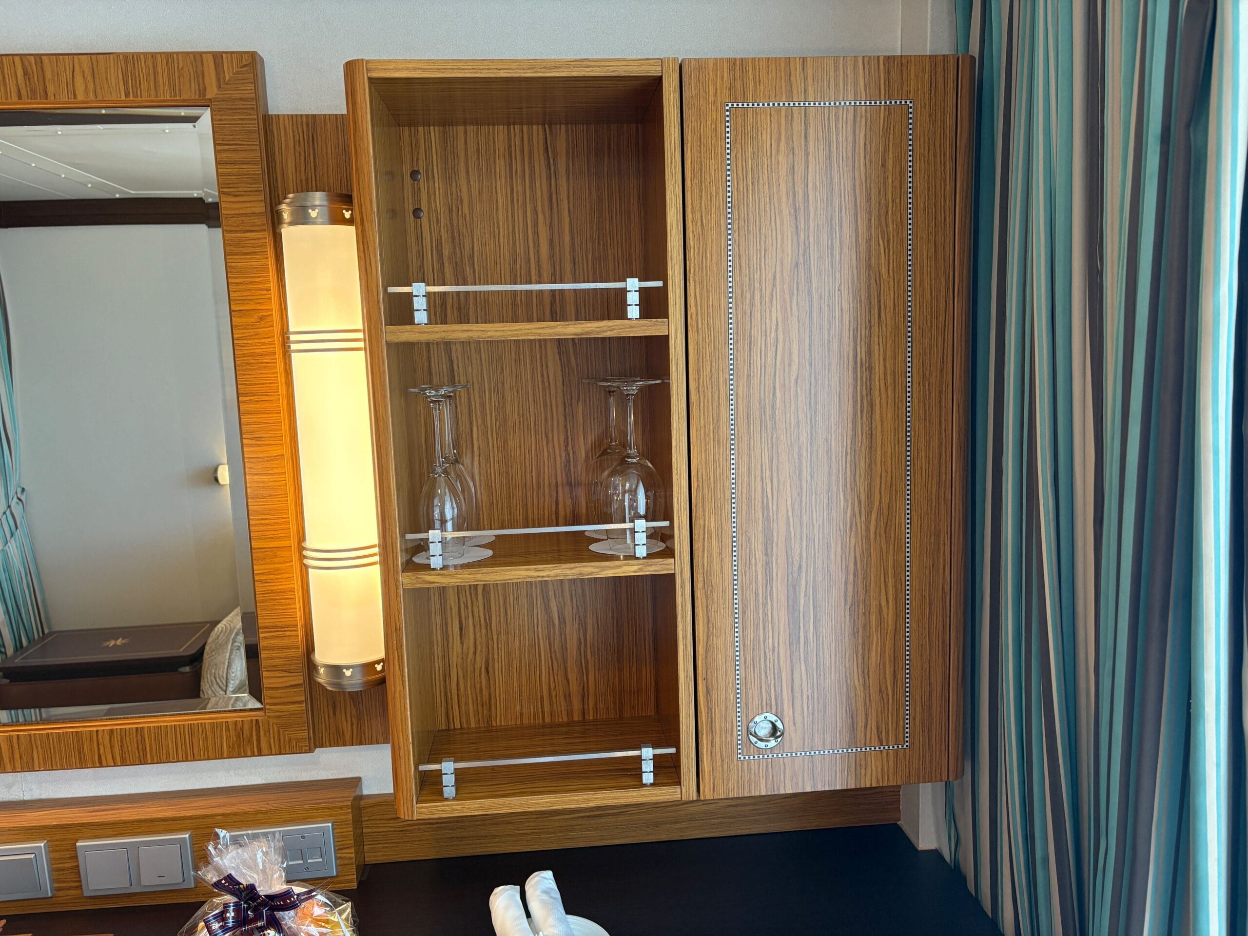 Open wooden cabinet with two wine glasses inside, beside a mirror, in a Disney Dream Concierge Family Oceanview Stateroom.
