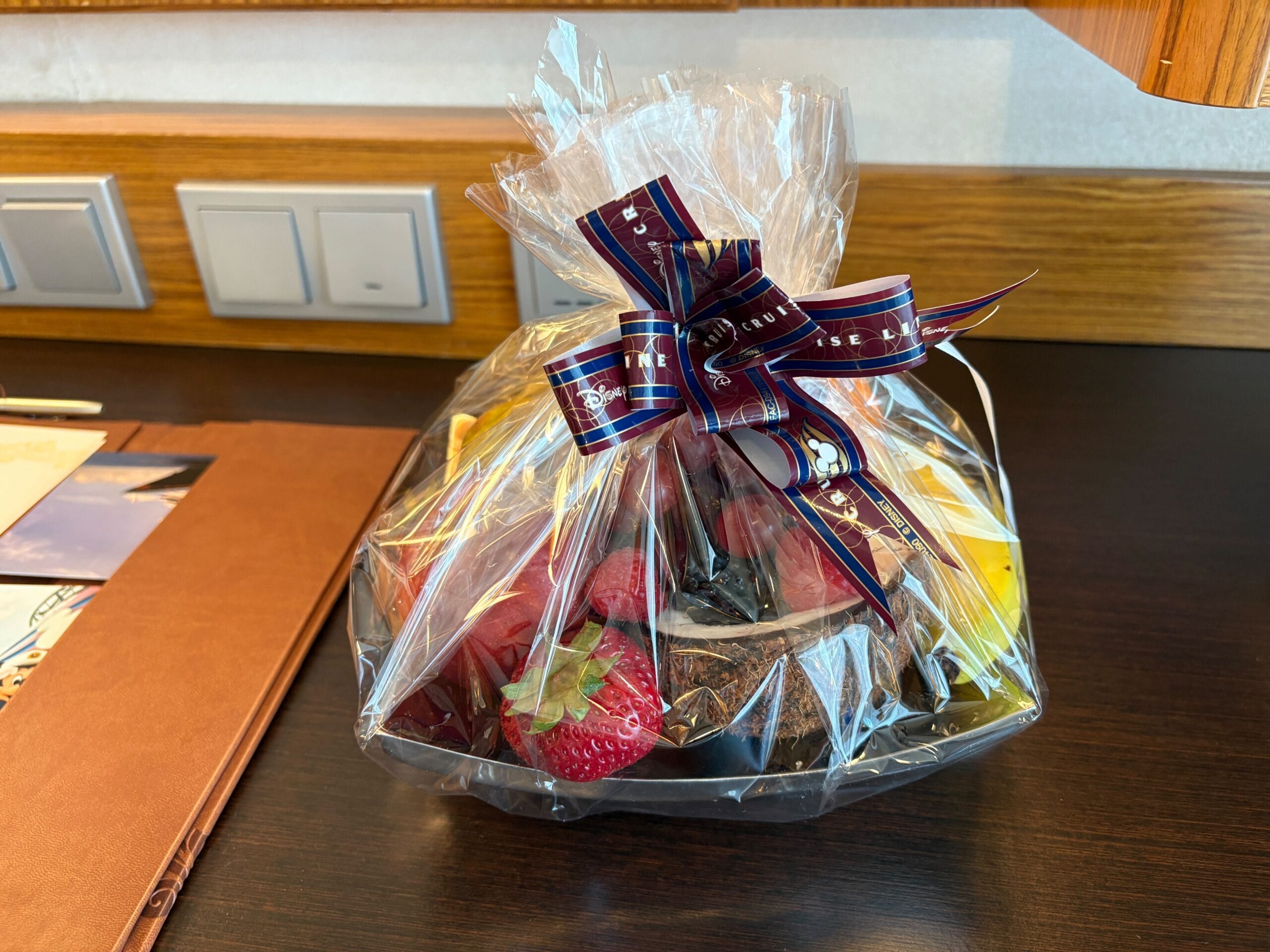 A gift basket of assorted fruit wrapped in clear plastic sits on a wooden desk in a Concierge Family Oceanview Stateroom.
