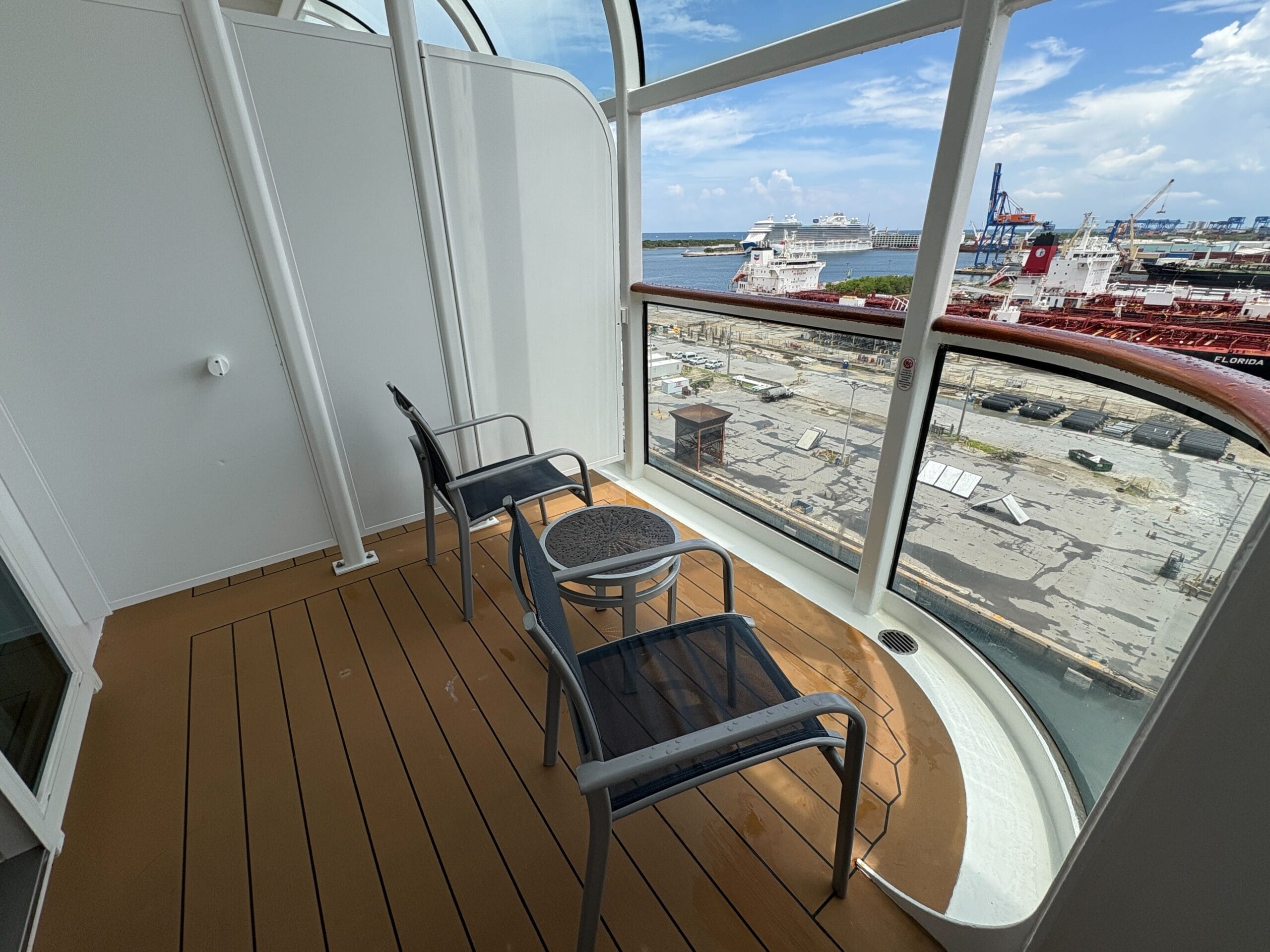 Two chairs and a small table on a Disney Dream balcony stateroom overlooking ships, port, and blue sky.