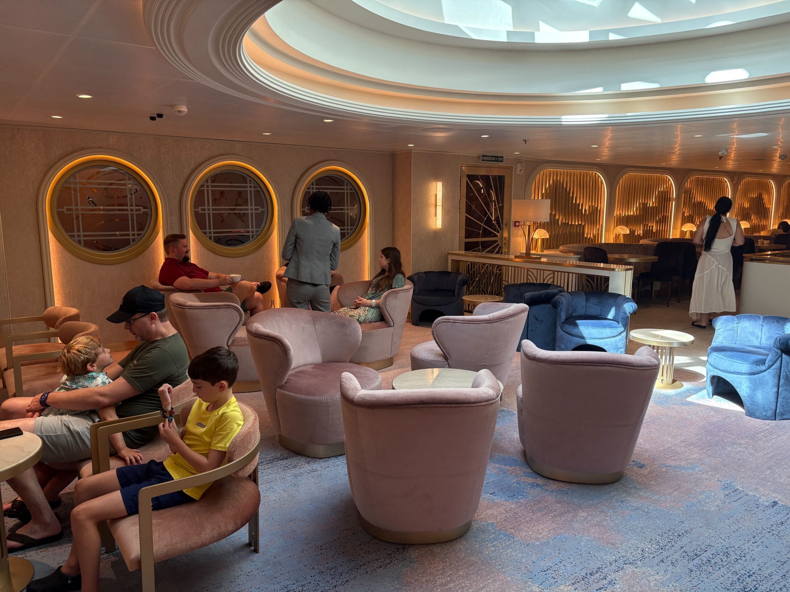 People relaxing in a lounge area on the Disney Dream, with plush chairs, round windows, and soft lighting.