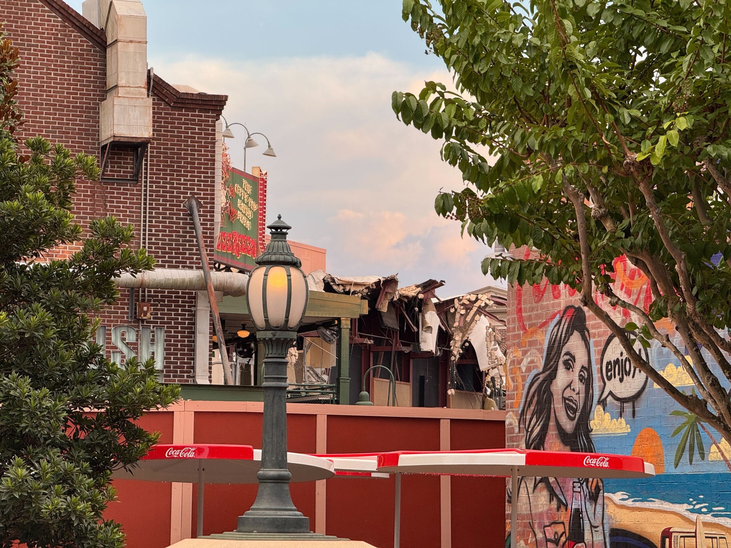 A demolished building with debris, beside a mural and streetlamp, behind red Coca-Cola barriers at Disney's Hollywood Studios.