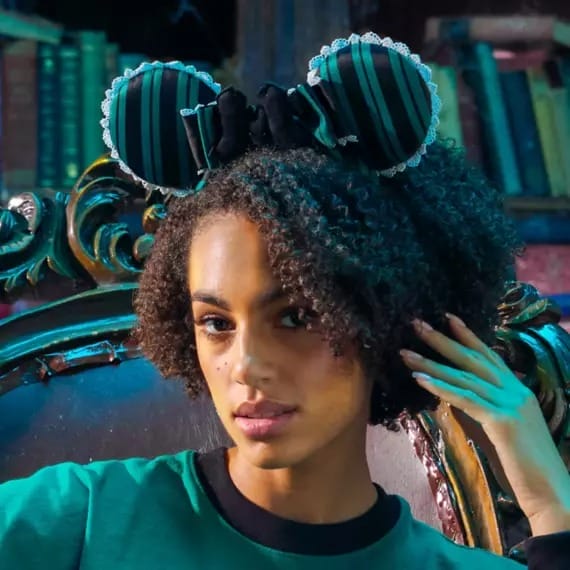 Woman with curly hair wears striped Minnie Mouse ears and a green shirt, sitting on an ornate chair in front of bookshelves.