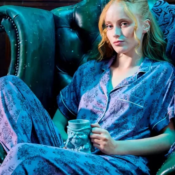 Woman in purple patterned pajamas sits on a green leather chair, holding a mug and looking at the camera.
