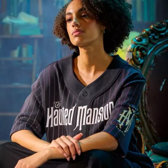 A woman sits on an ornate chair wearing a black "Haunted Mansion" shirt in a dimly lit, mysterious room.