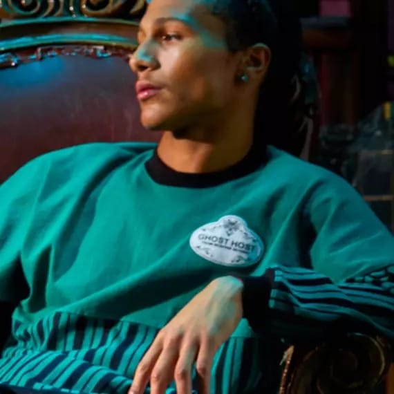 A person in a teal sweater with a "Ghost Host" patch sits on a dark ornate chair, looking to the side.