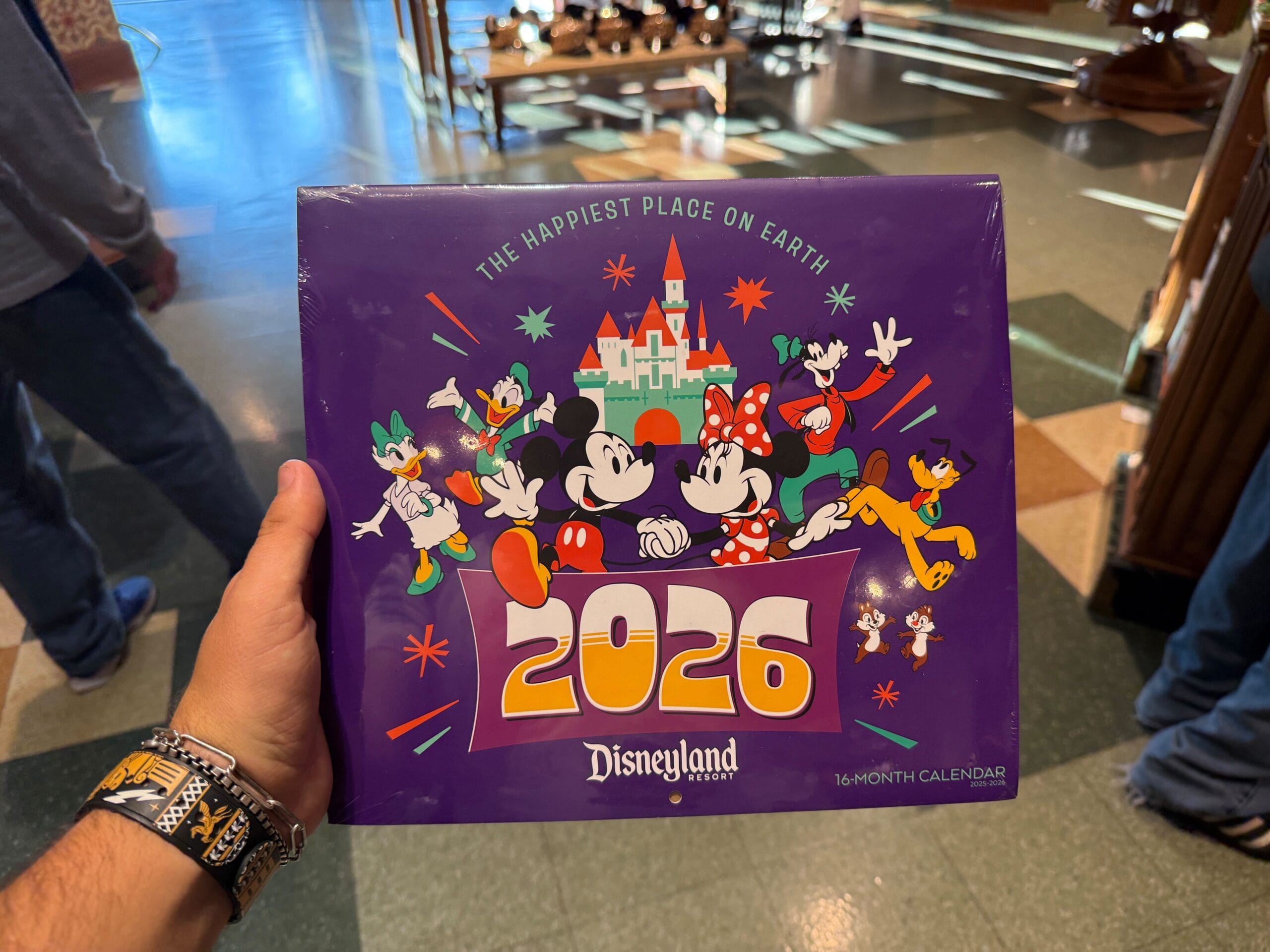 A hand holds a Disneyland 2026 calendar with Mickey, Minnie, and other characters on a purple cover.
