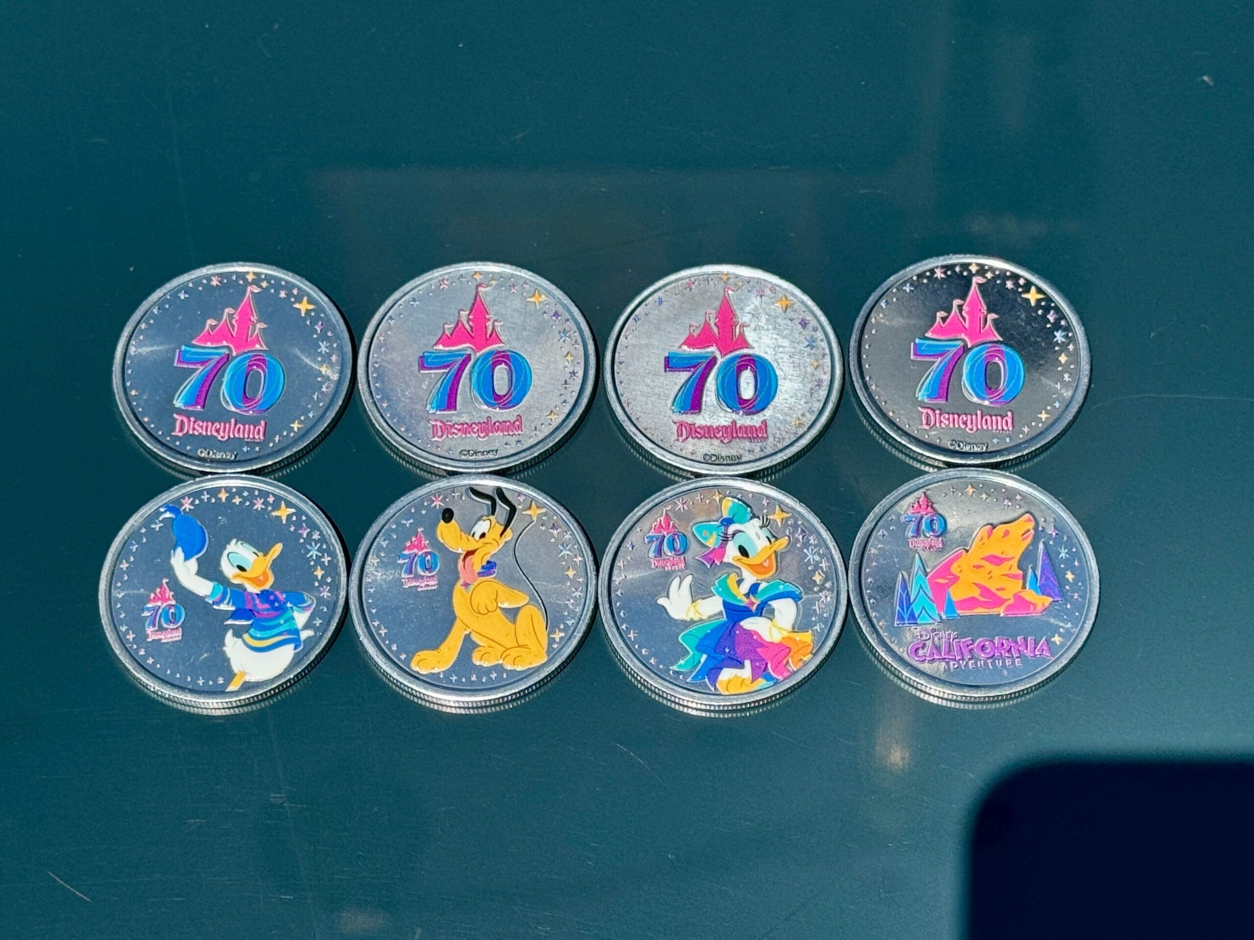 Eight Disneyland 70th Anniversary collectible medallions feature Disney characters and vibrant designs on a dark surface.