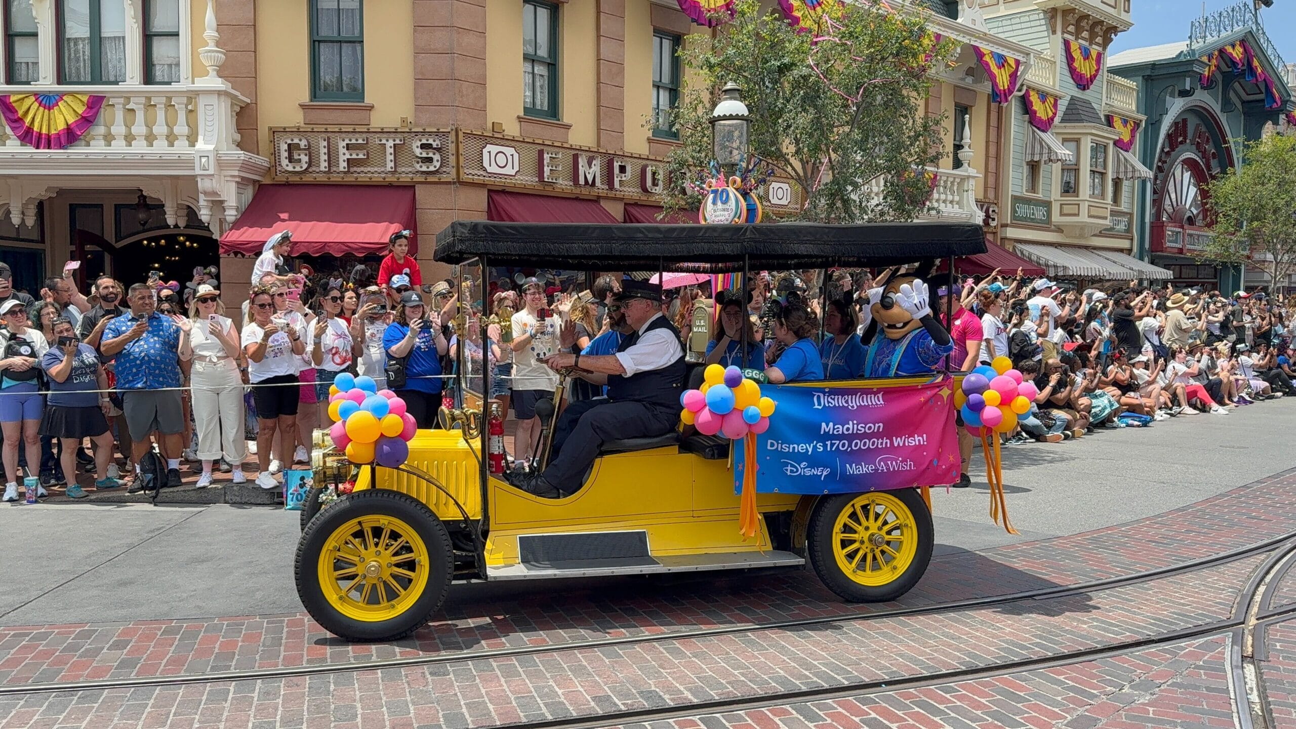 A yellow vintage car decorated with balloons leads a Disneyland birthday cavalcade as crowds cheer along the street.