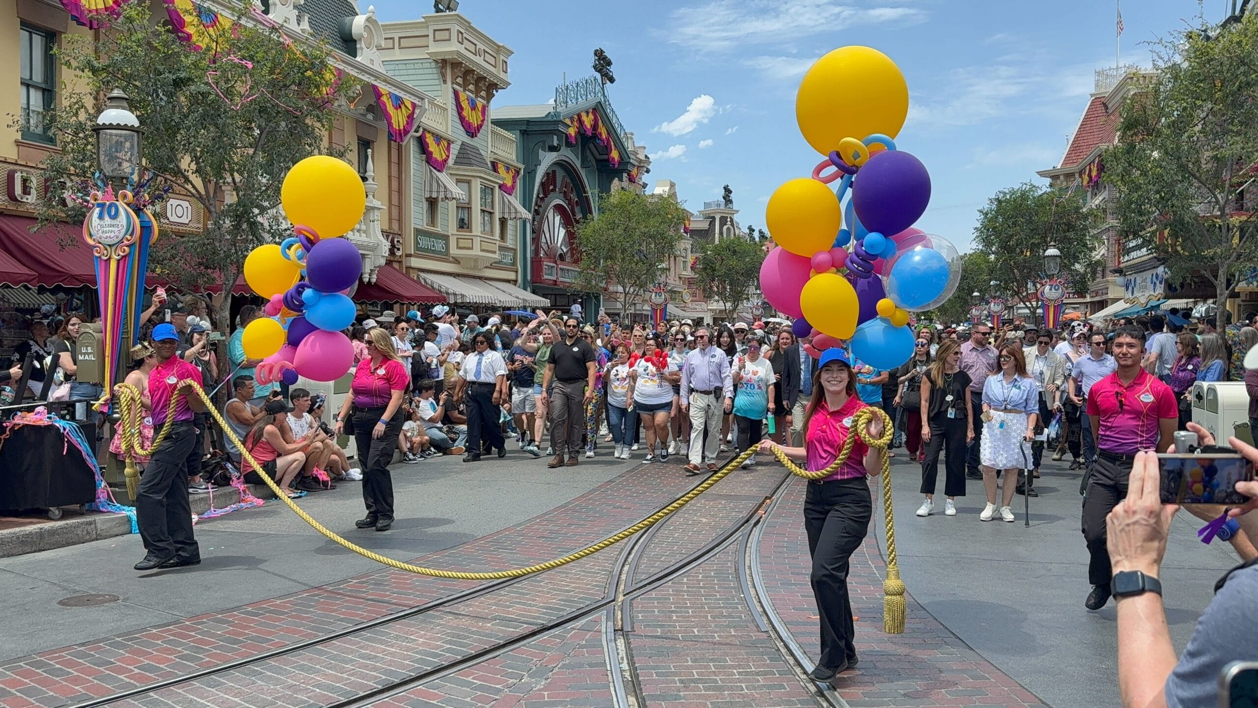 Performers in colorful uniforms join the Disneyland birthday cavalcade as crowds watch on a lively, festive street.