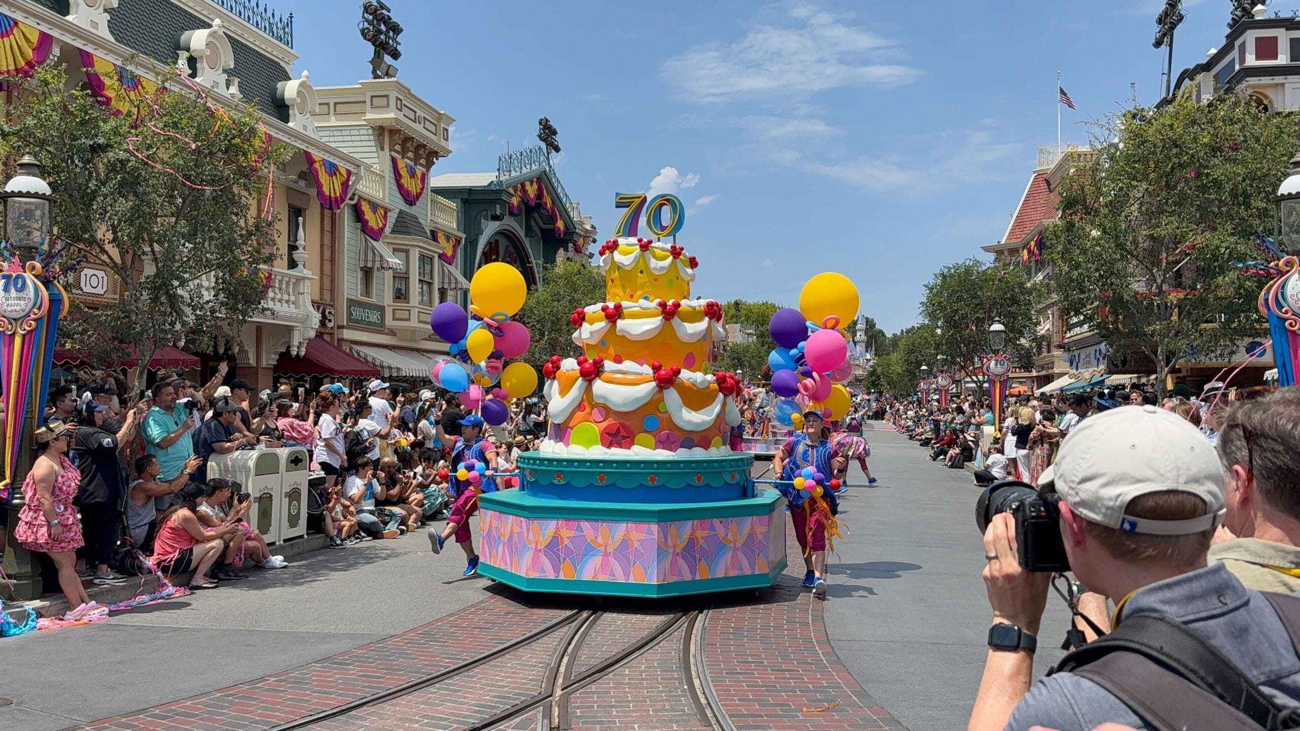 A colorful Disneyland birthday cavalcade float shaped like a cake with "70" on top moves down a theme park street.
