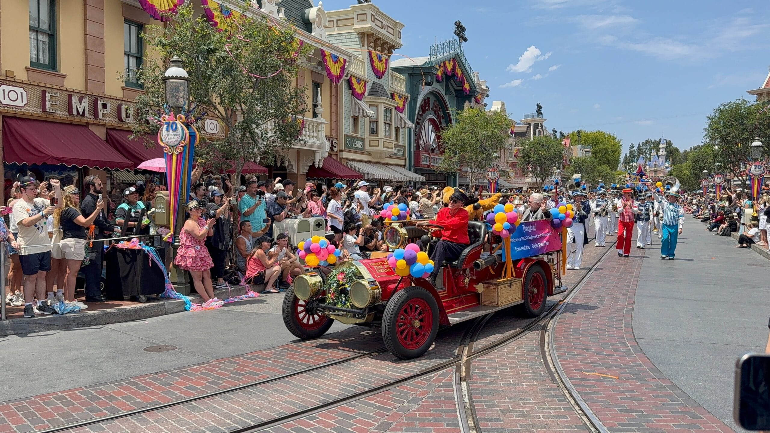 A festive disneyland birthday cavalcade with people on a vintage fire truck, cheered by a crowd on a sunny day.