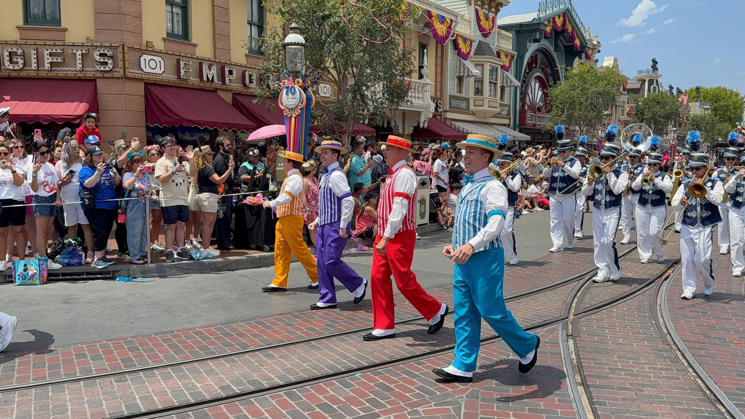 A colorful Disneyland Birthday Cavalcade with costumed performers and a marching band delights crowds on a brick street.