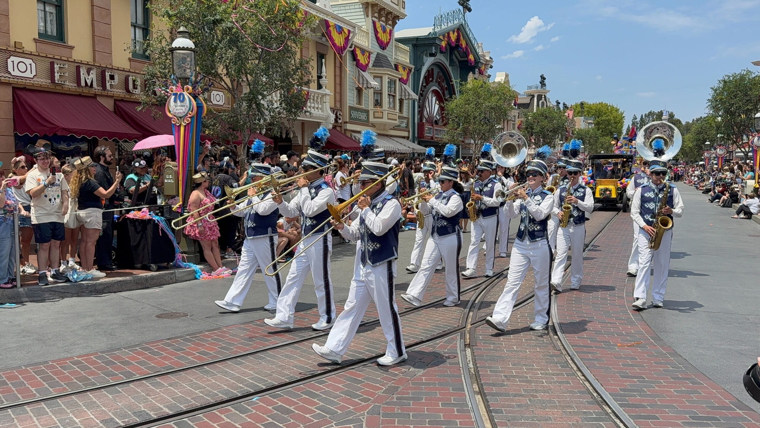 A marching band in white uniforms plays in the Disneyland Birthday Cavalcade as spectators watch the lively, decorated street.