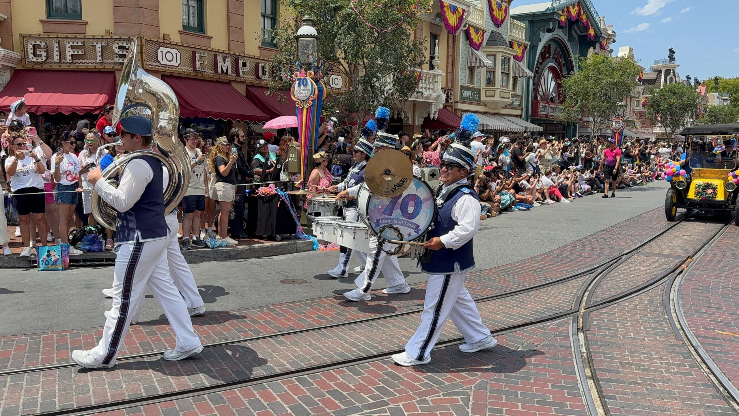 A marching band in blue and white uniforms performs in a Disneyland birthday cavalcade as crowds watch from the sidewalk.