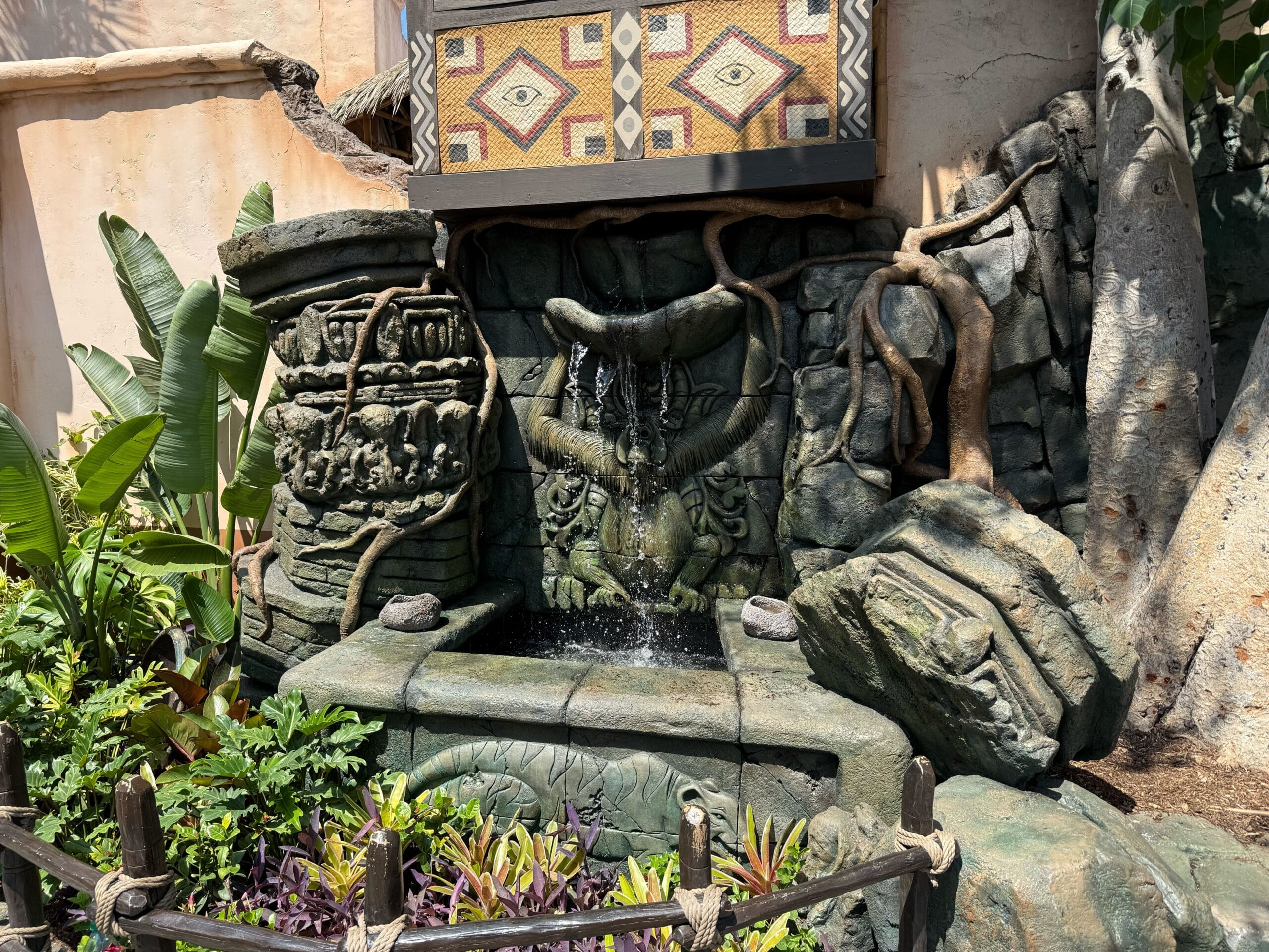 Decorative stone fountain with intricate carvings, surrounded by plants and a patterned tile mural above.