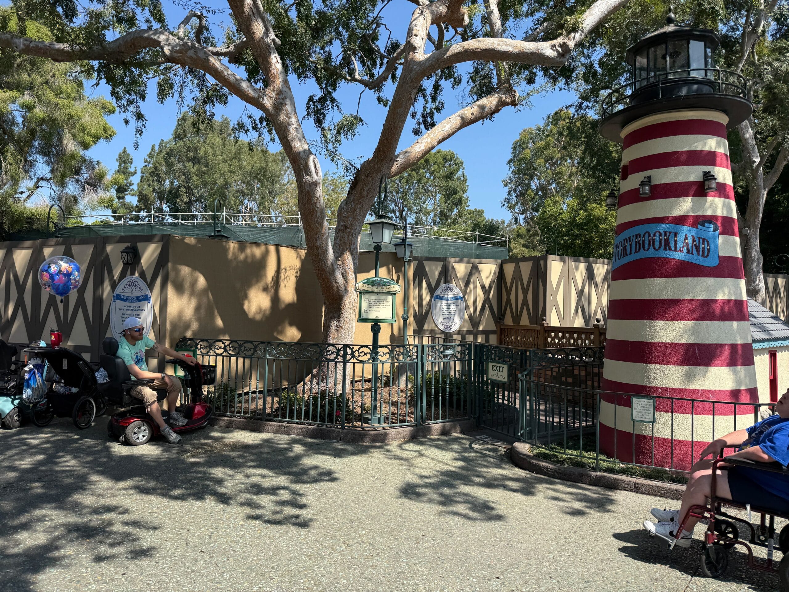 Entrance to Storybook Land with a striped lighthouse, tree, strollers, and people sitting in the shade.