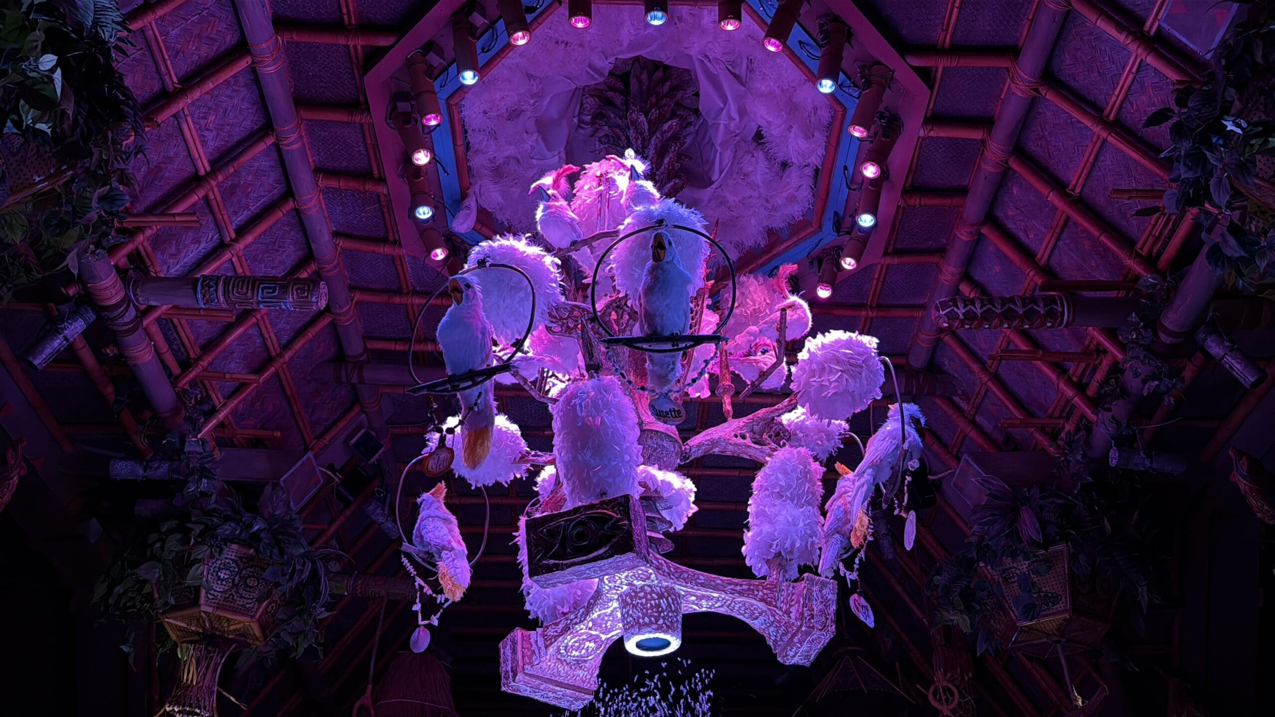 A brightly lit chandelier with animatronic birds, glowing in purple and pink lights, hangs from a bamboo ceiling.