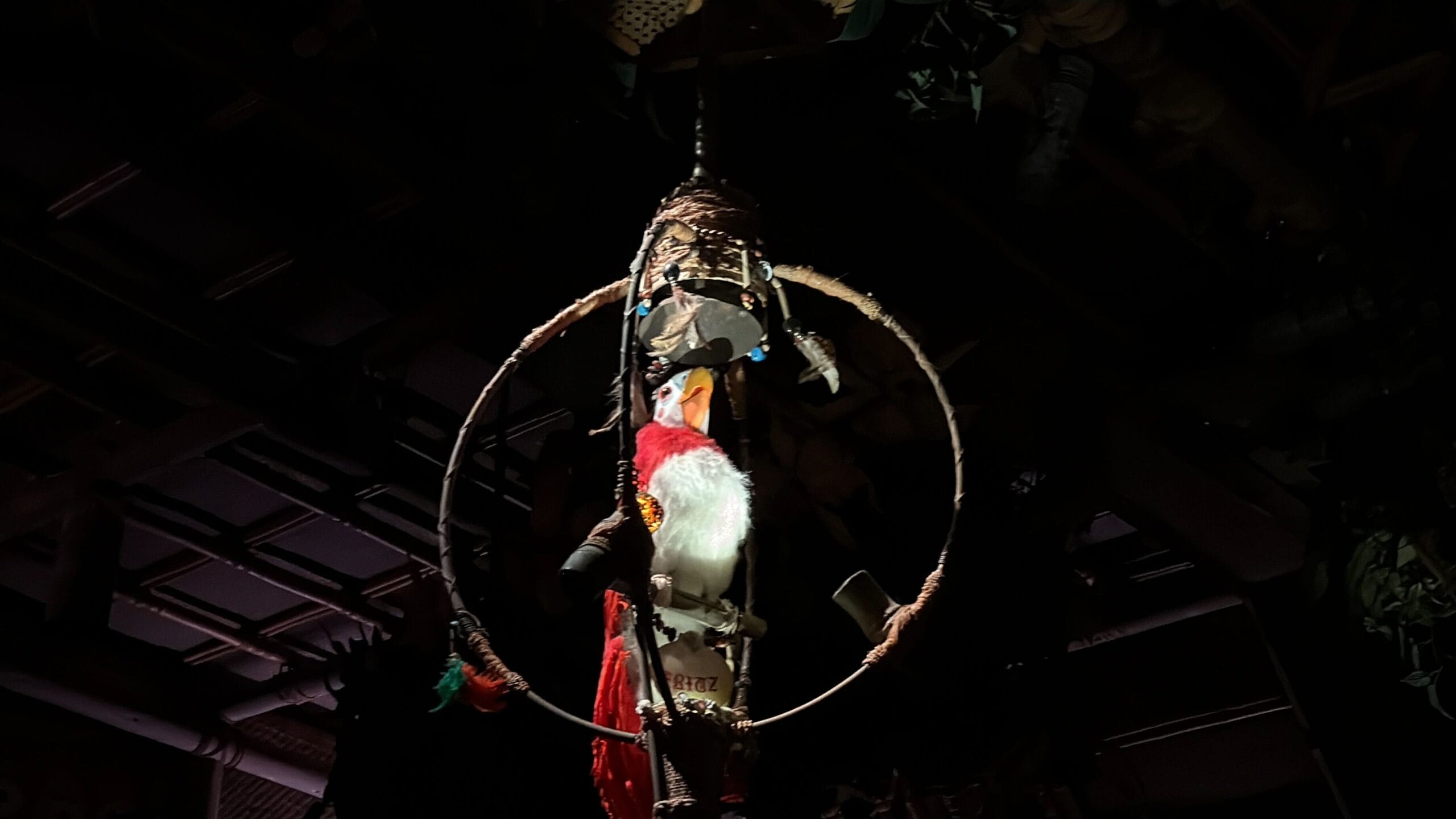 A colorful animatronic parrot sits on a wooden perch, illuminated by a spotlight in a dark room.