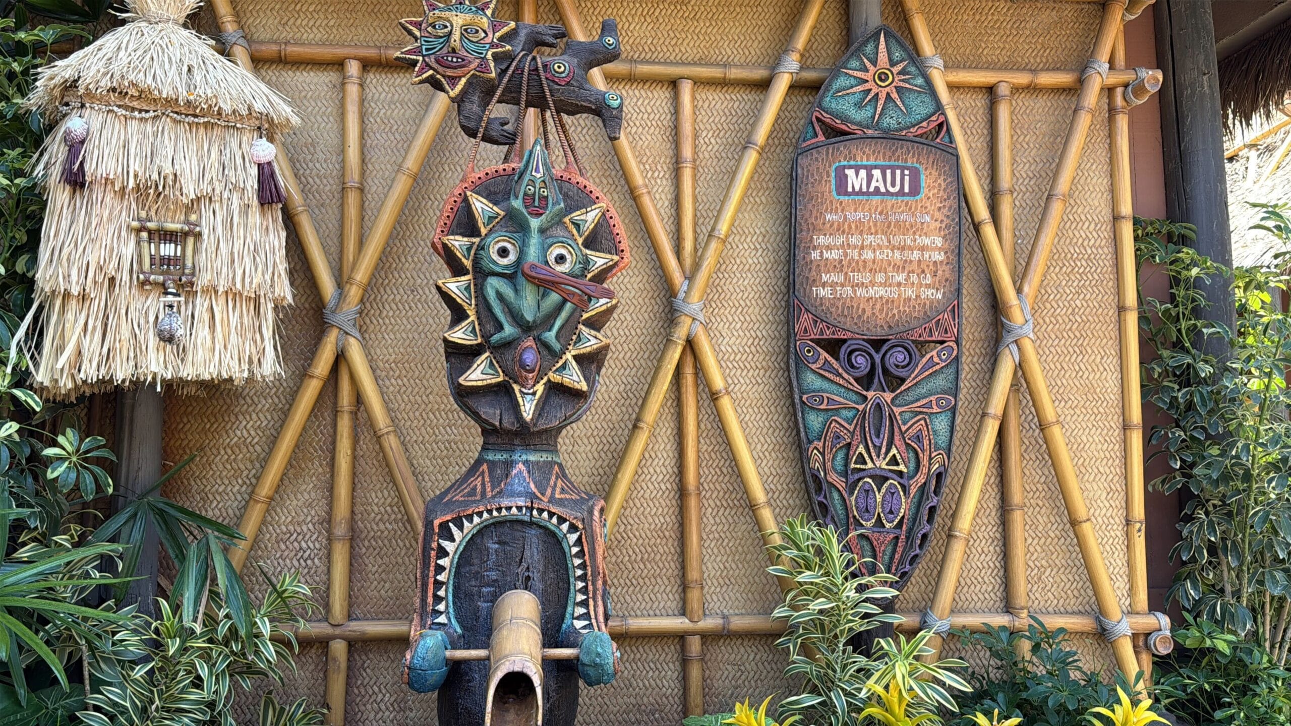Colorful tiki statues and a "Maui" shield on bamboo at Disneyland's Enchanted Tiki Room reopening, with plants and lanterns.