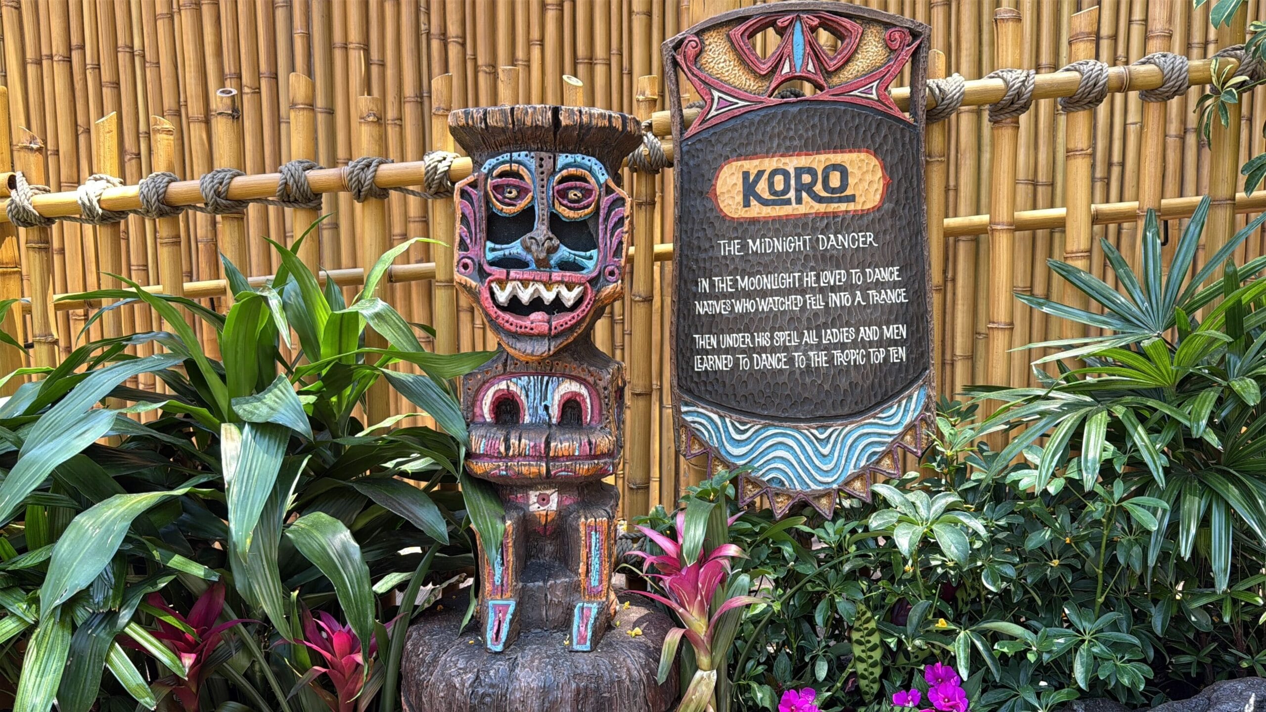 Colorful tiki statue and "Koro" sign by bamboo evoke Disneyland’s Enchanted Tiki Room in a lush outdoor setting.
