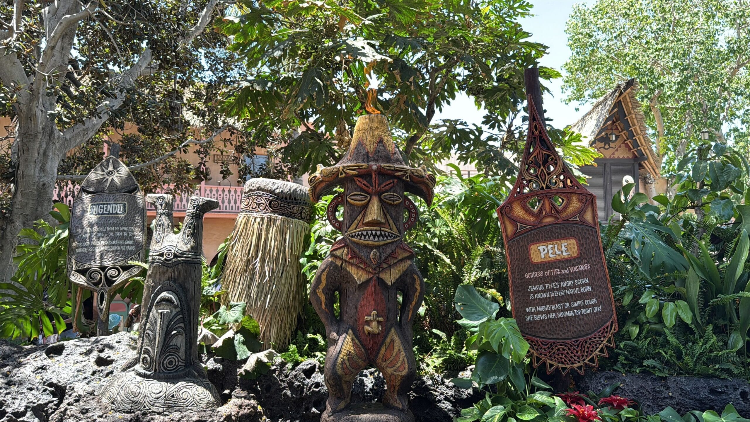 Four tiki statues and plaques in lush greenery, with trees and Disneyland’s enchanted Tiki Room in the background.