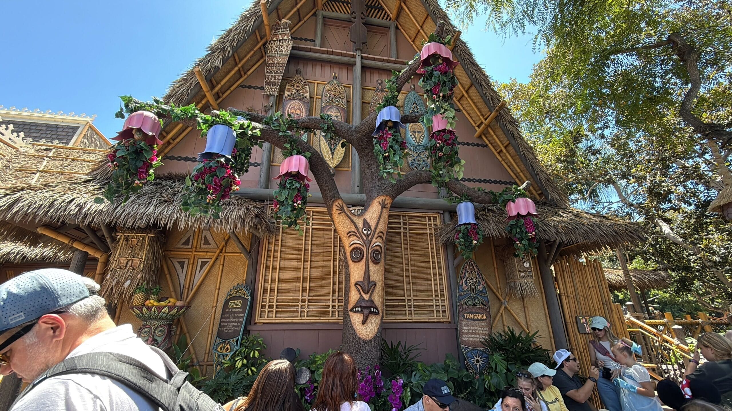A whimsical tiki hut near a carved face tree, as crowds gather for the enchanted tiki room reopening in Disneyland.