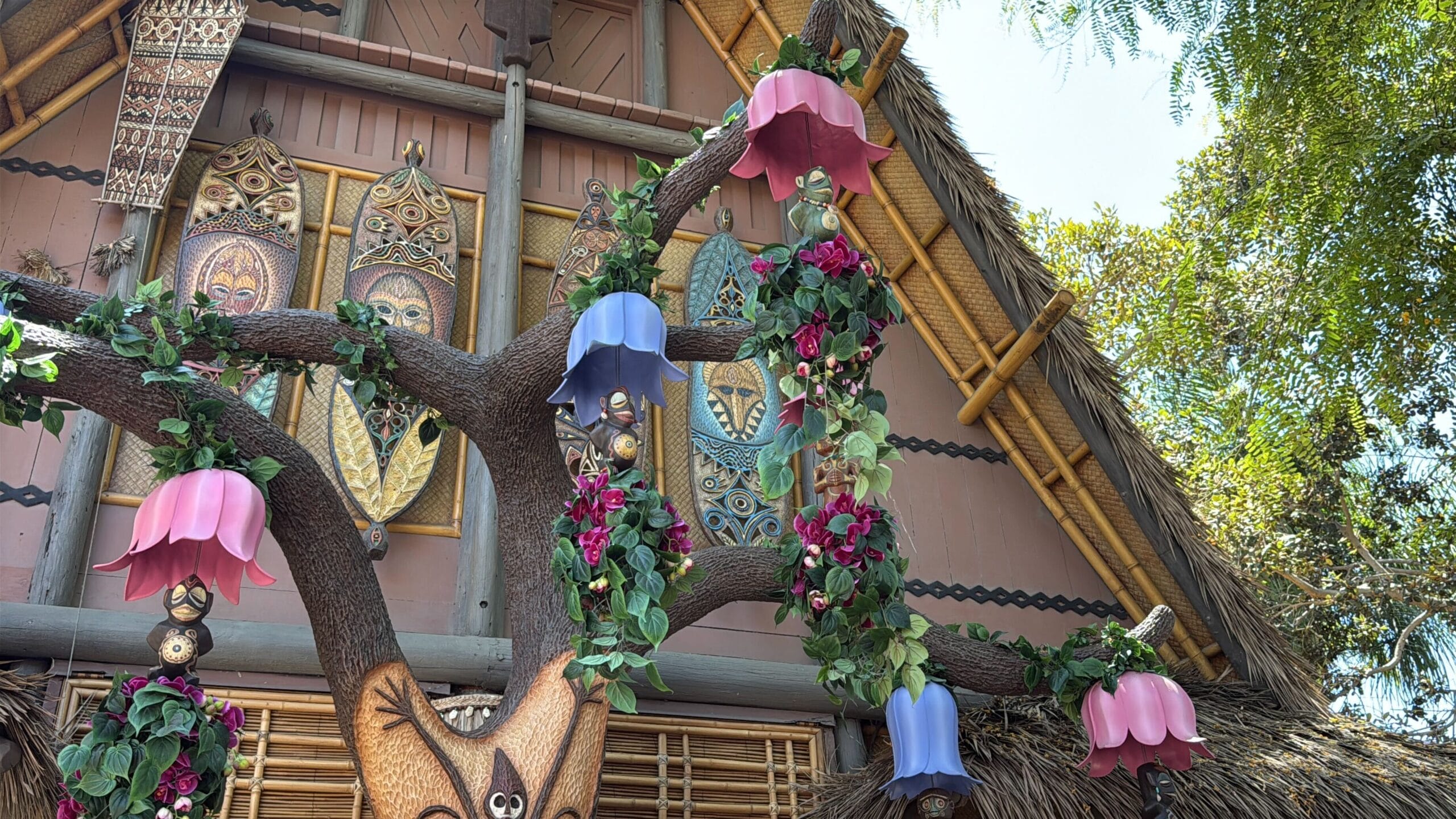 Colorful lanterns and flowers hang from a tree near the Disneyland Enchanted Tiki Room with tribal masks and decor.