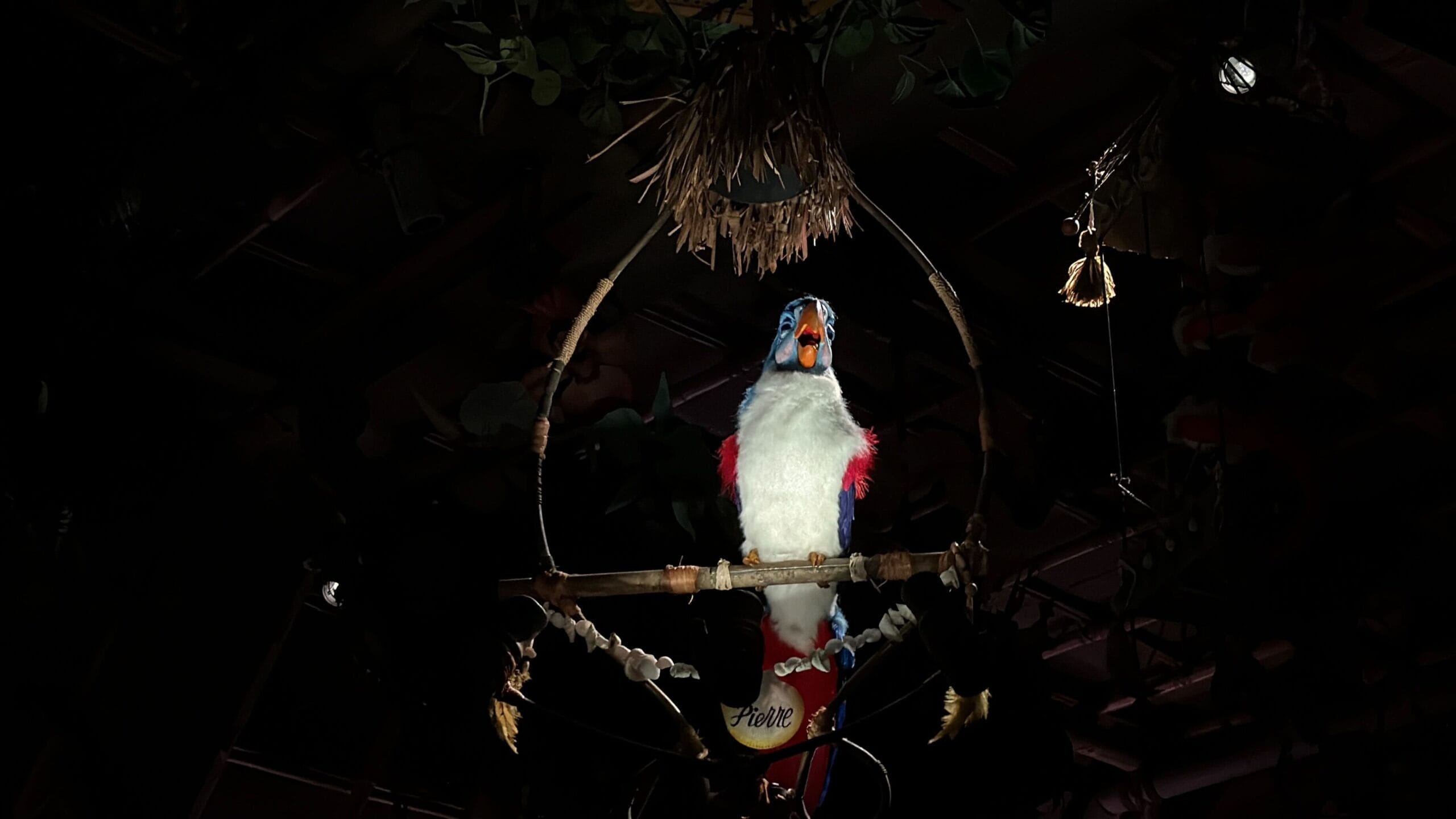 A colorful animatronic parrot sits on a perch under a spotlight, with a dark background.