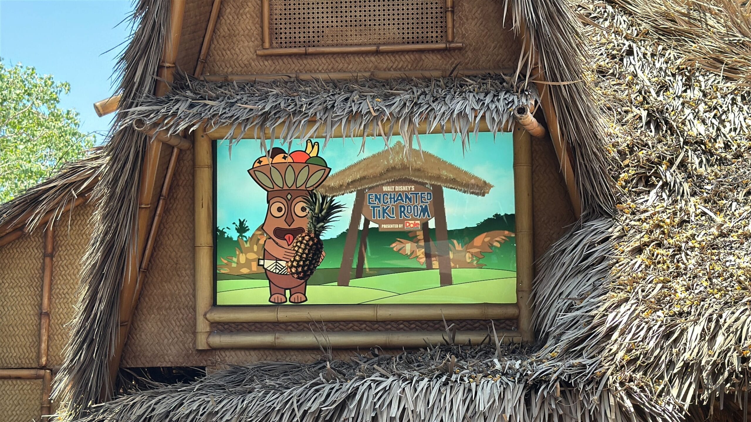 A colorful Enchanted Tiki Room sign marks its reopening at Disneyland, nestled among lush tropical foliage.