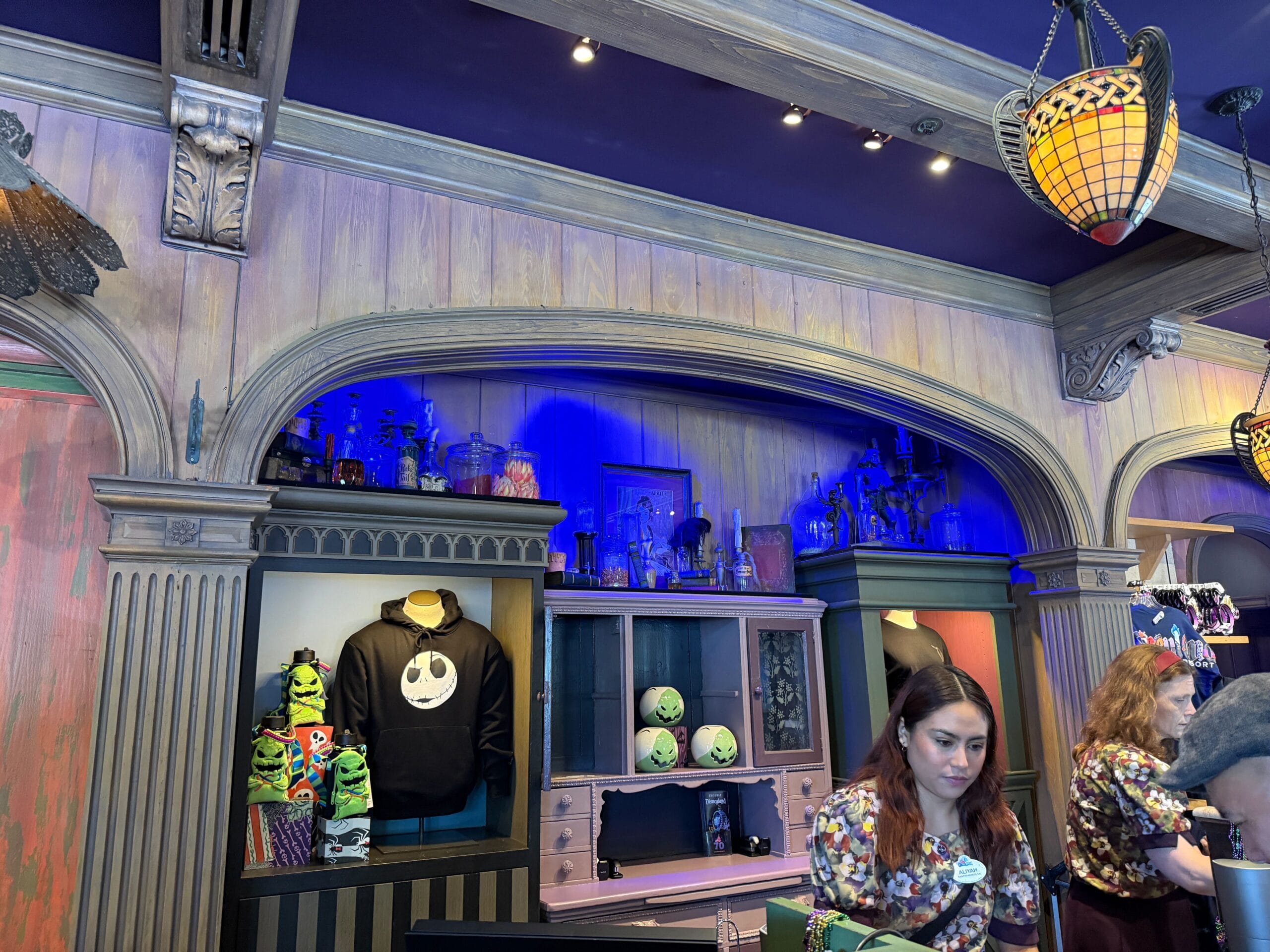 Two employees stand at a counter in a themed shop selling Haunted Mansion and Nightmare Before Christmas merchandise.